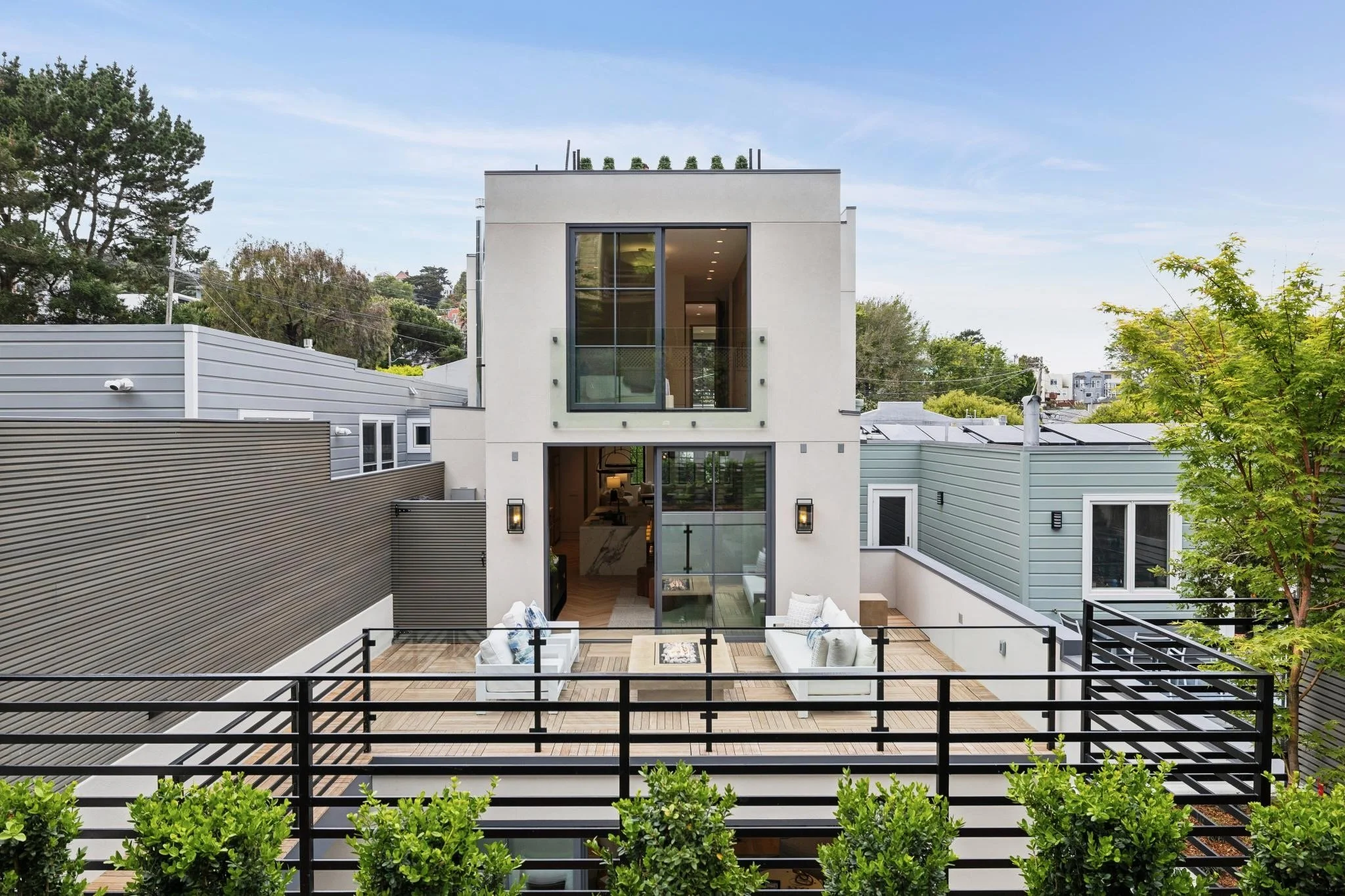 Modern multi-story house with large windows, white exterior, rooftop garden, and a patio with outdoor seating and potted plants.