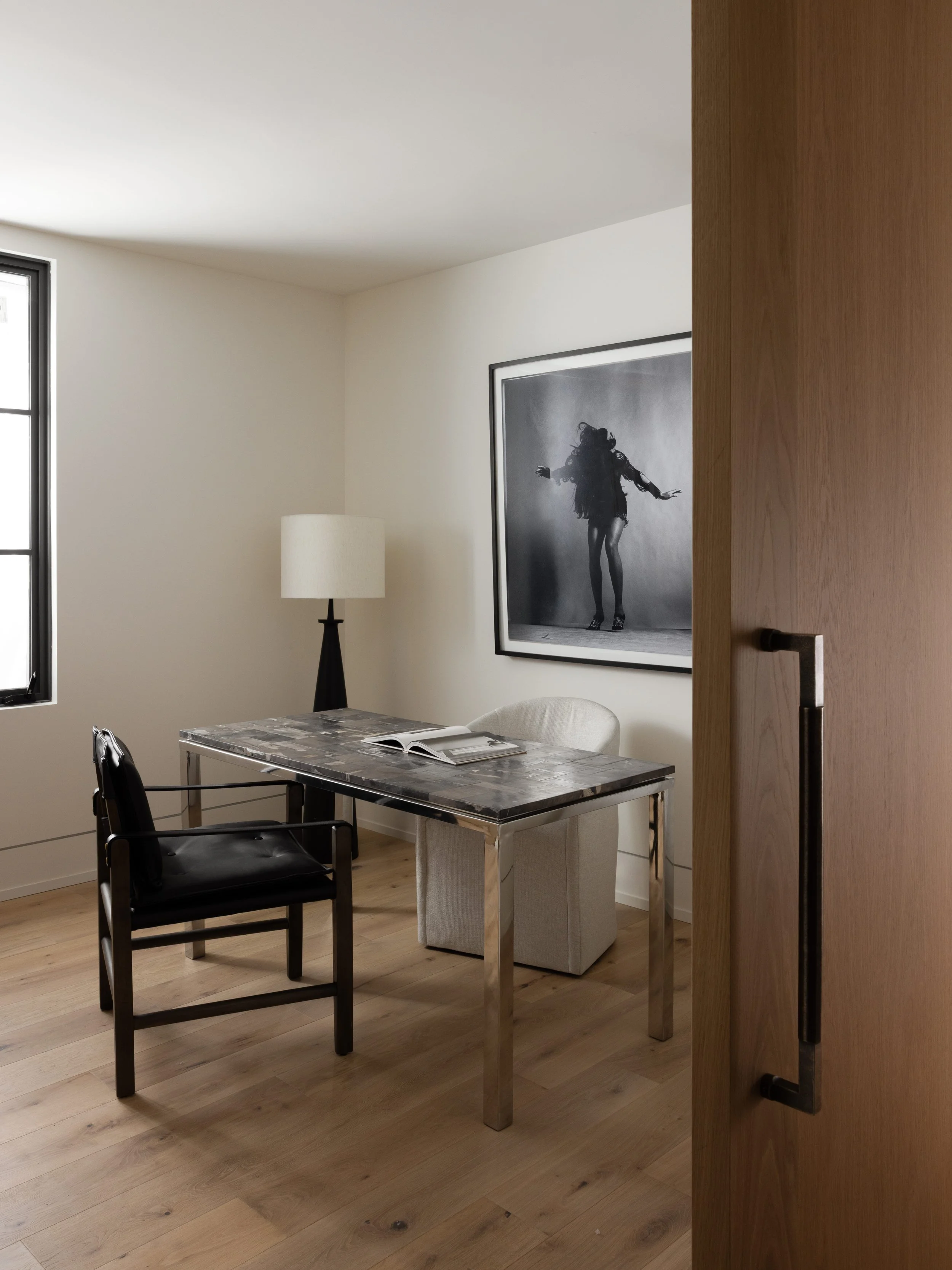 Interior of a modern room with a black and white photo on the wall, a table with an open magazine, two chairs, a window, and a floor lamp.