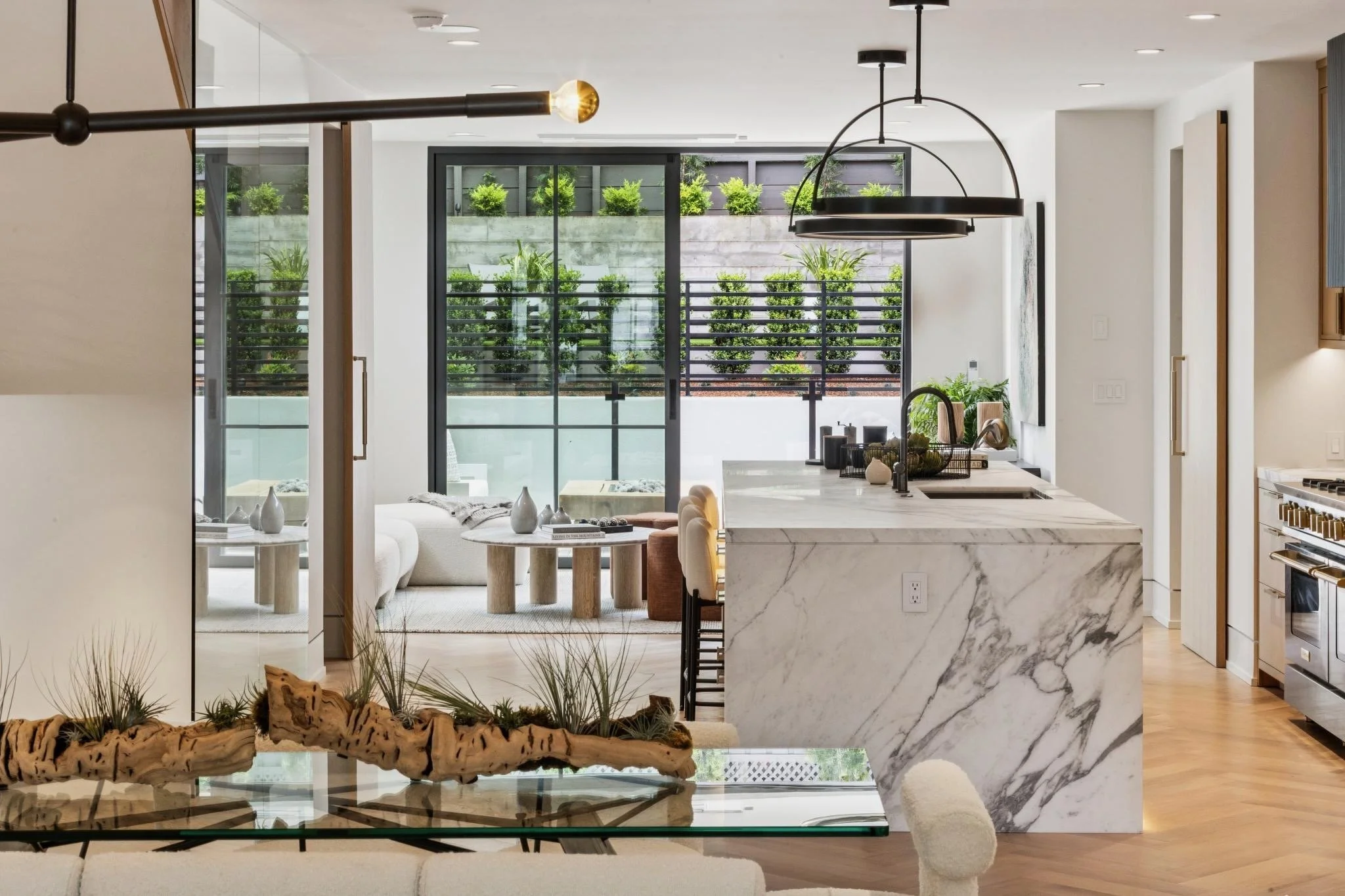 Interior view of a modern kitchen and living space with white walls, a marble island, black lighting fixtures, and large sliding glass doors leading to an outdoor area with green plants.