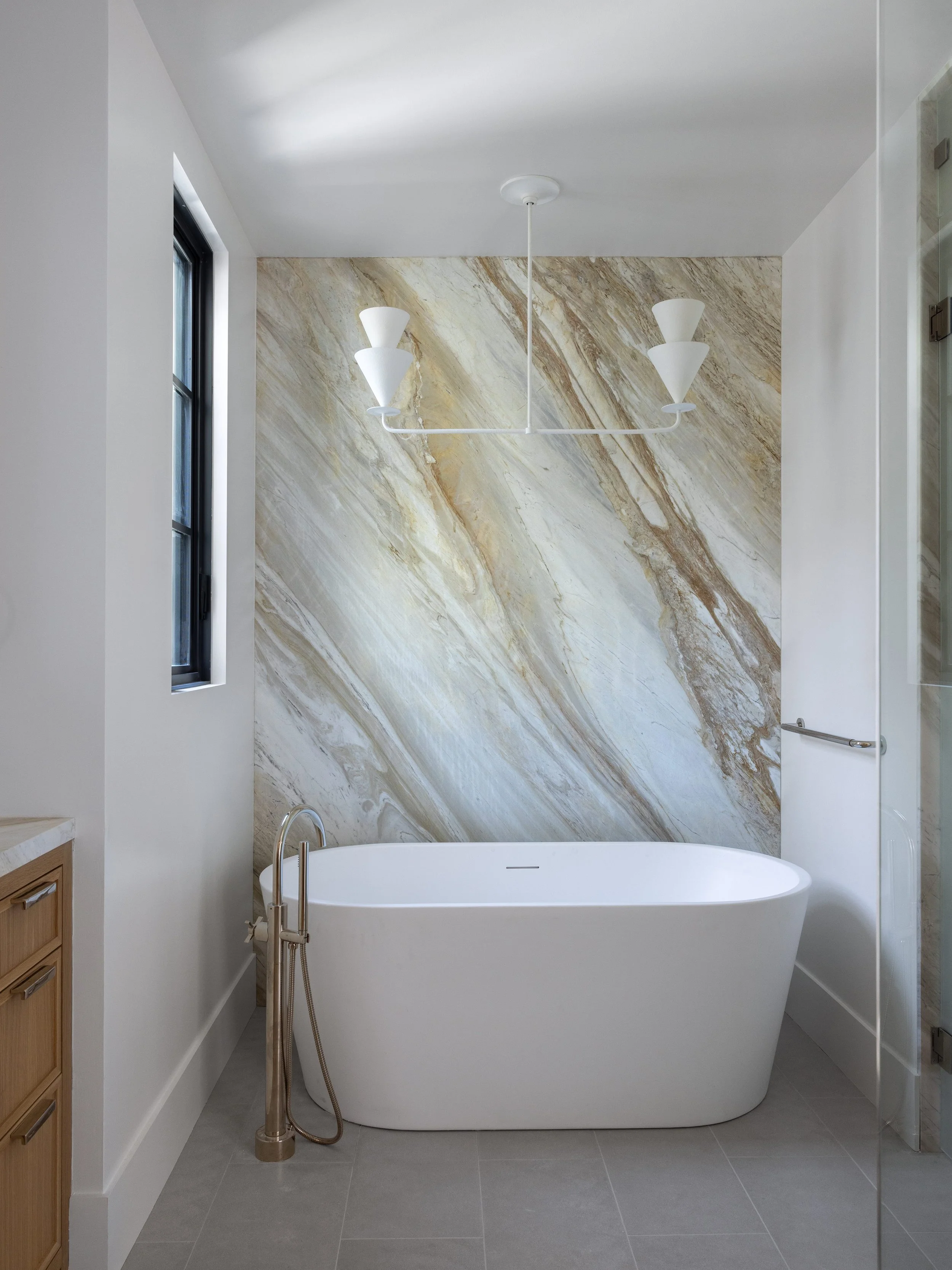 Modern bathroom with a white freestanding bathtub against a marble accent wall, a window on the left, a chandelier hanging from the ceiling, and part of a wooden cabinet on the left.