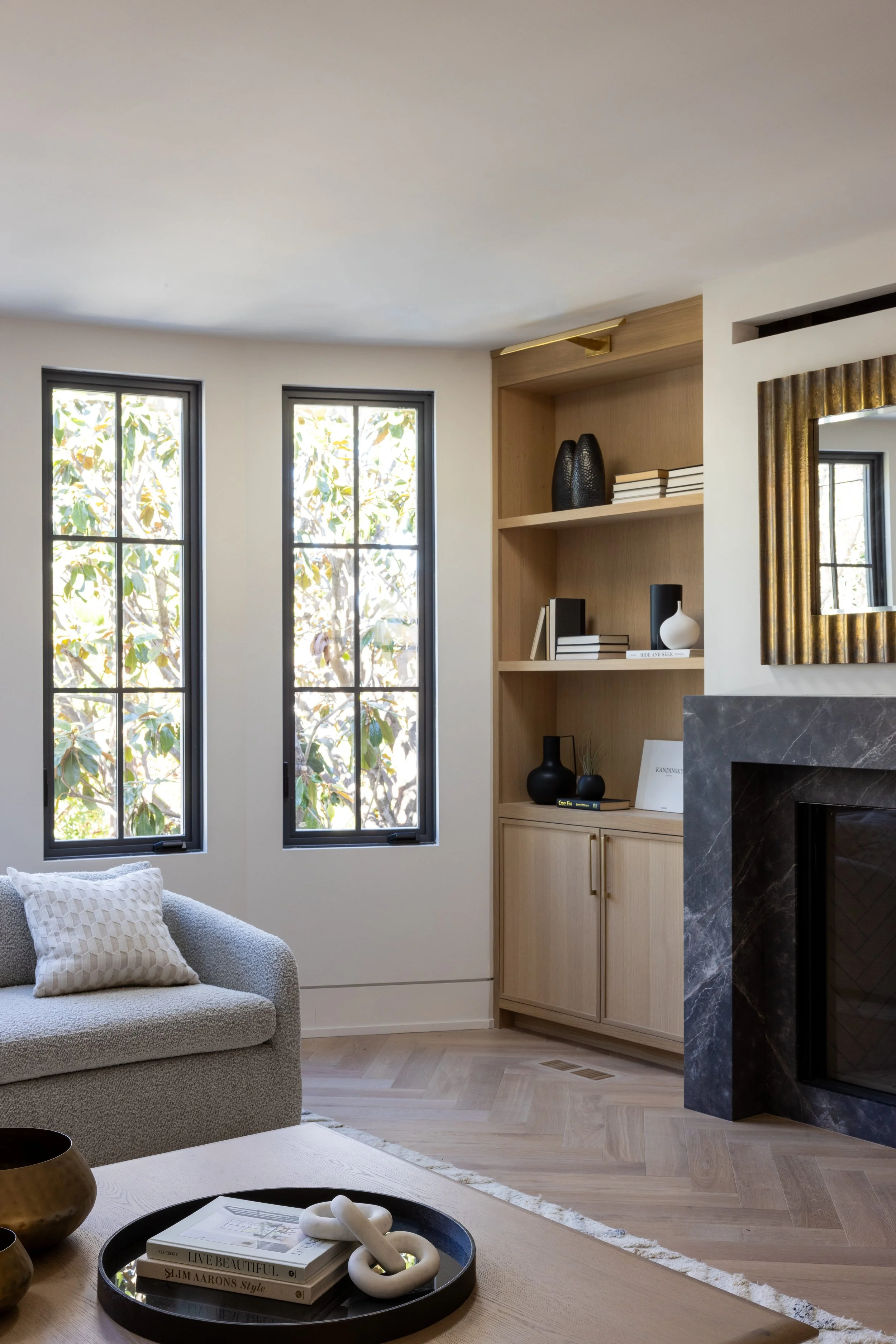 Modern living room with two large windows, a beige sofa with a textured pillow, a black and gold coffee table with books, a built-in wooden bookshelf with decorative vases and books, a black marble fireplace, and a gold framed mirror.