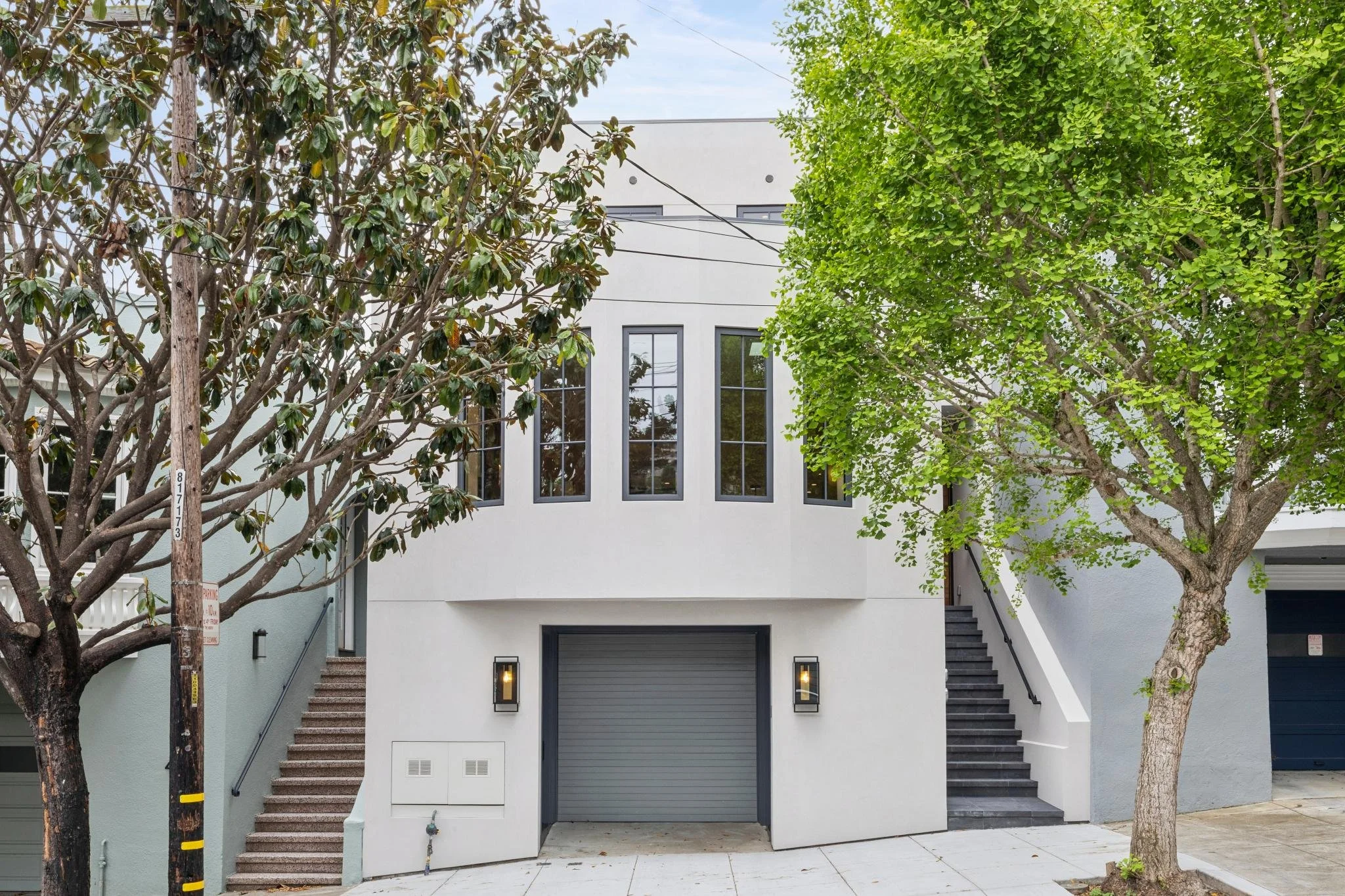 Modern white multi-story building with large windows and garage, flanked by trees on a city street.