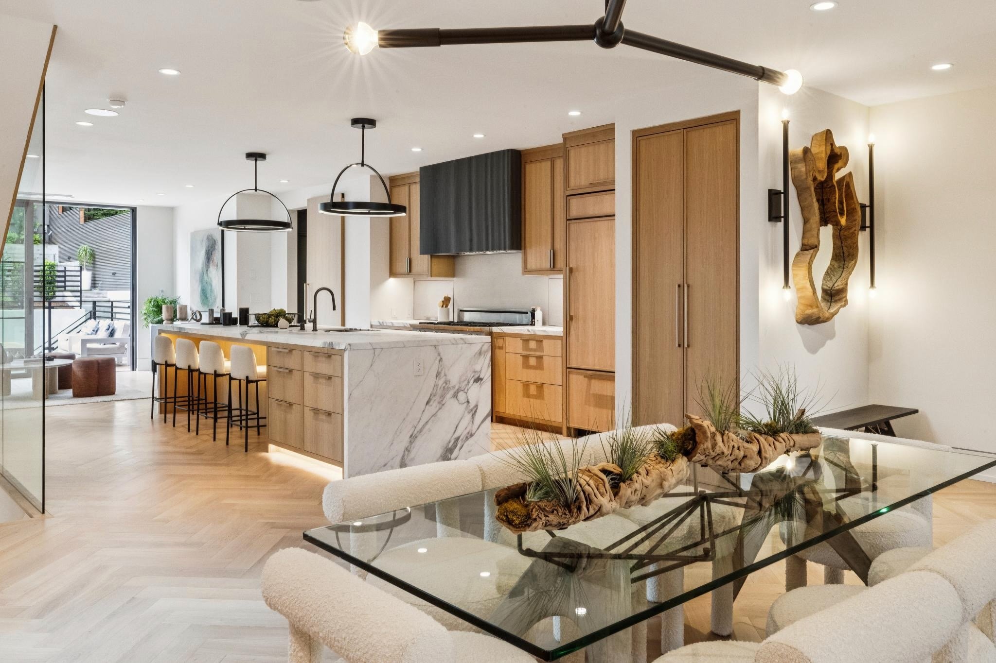 Modern open-concept kitchen and living area with wood cabinets, marble island, black pendant lights, and a nature-inspired decor with driftwood and plants.
