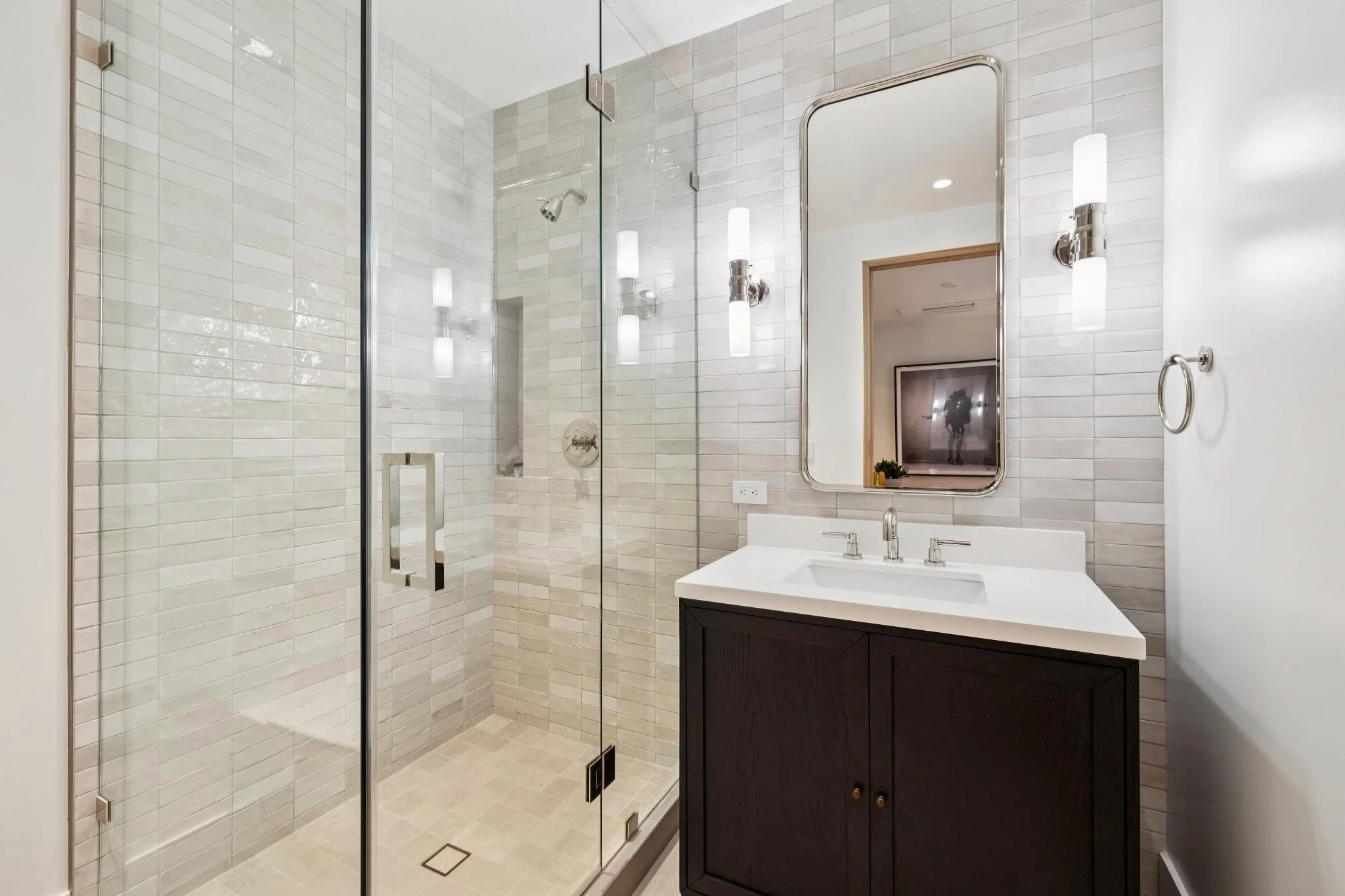 Modern bathroom with a glass-enclosed shower featuring white subway tiles, a small vanity with a dark cabinet, a white countertop, a rectangular mirror, and two wall sconces.
