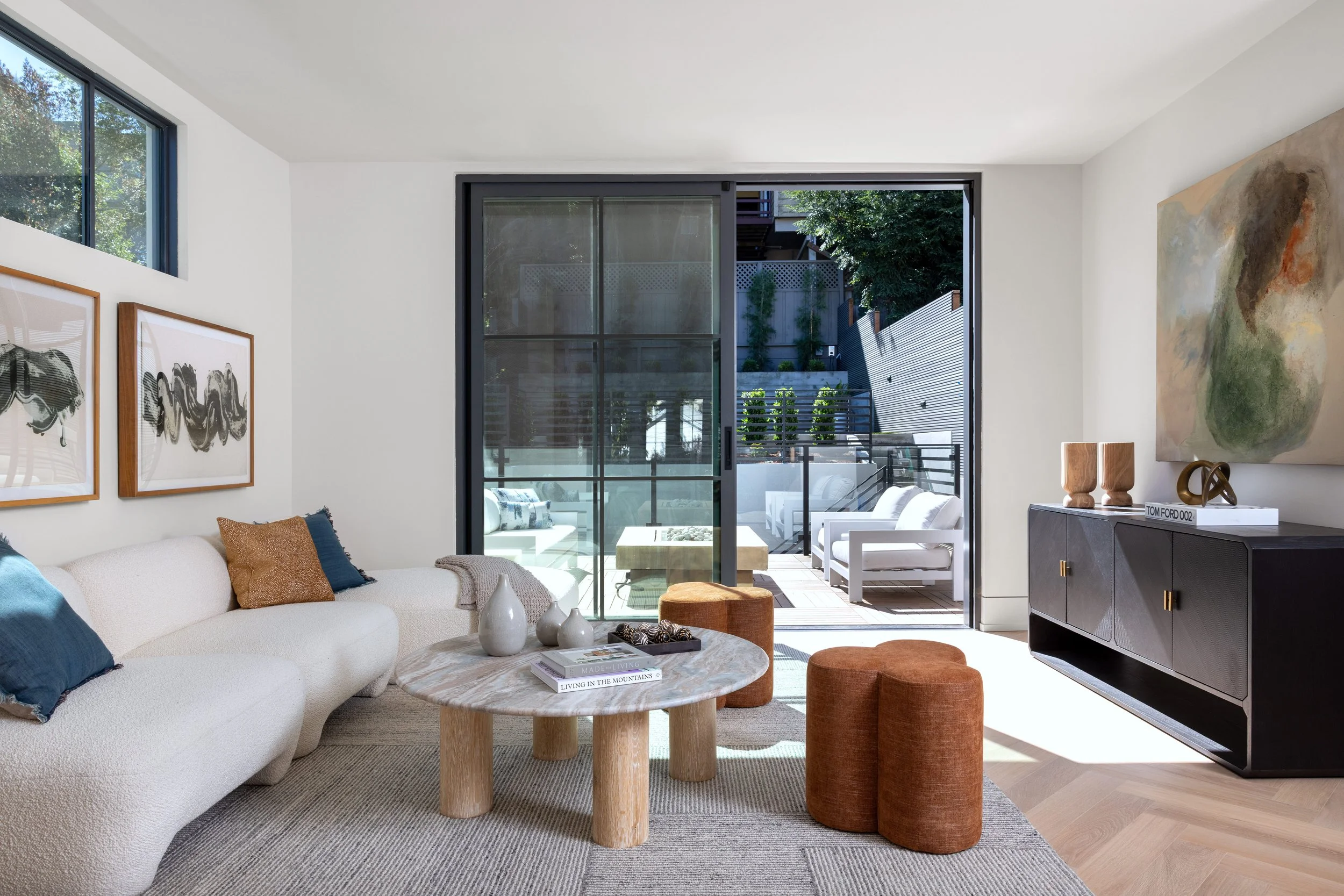 Modern living room with white sofa, abstract art on the walls, round marble coffee table, two orange poufs, and a balcony with outdoor seating and greenery visible through sliding glass doors.