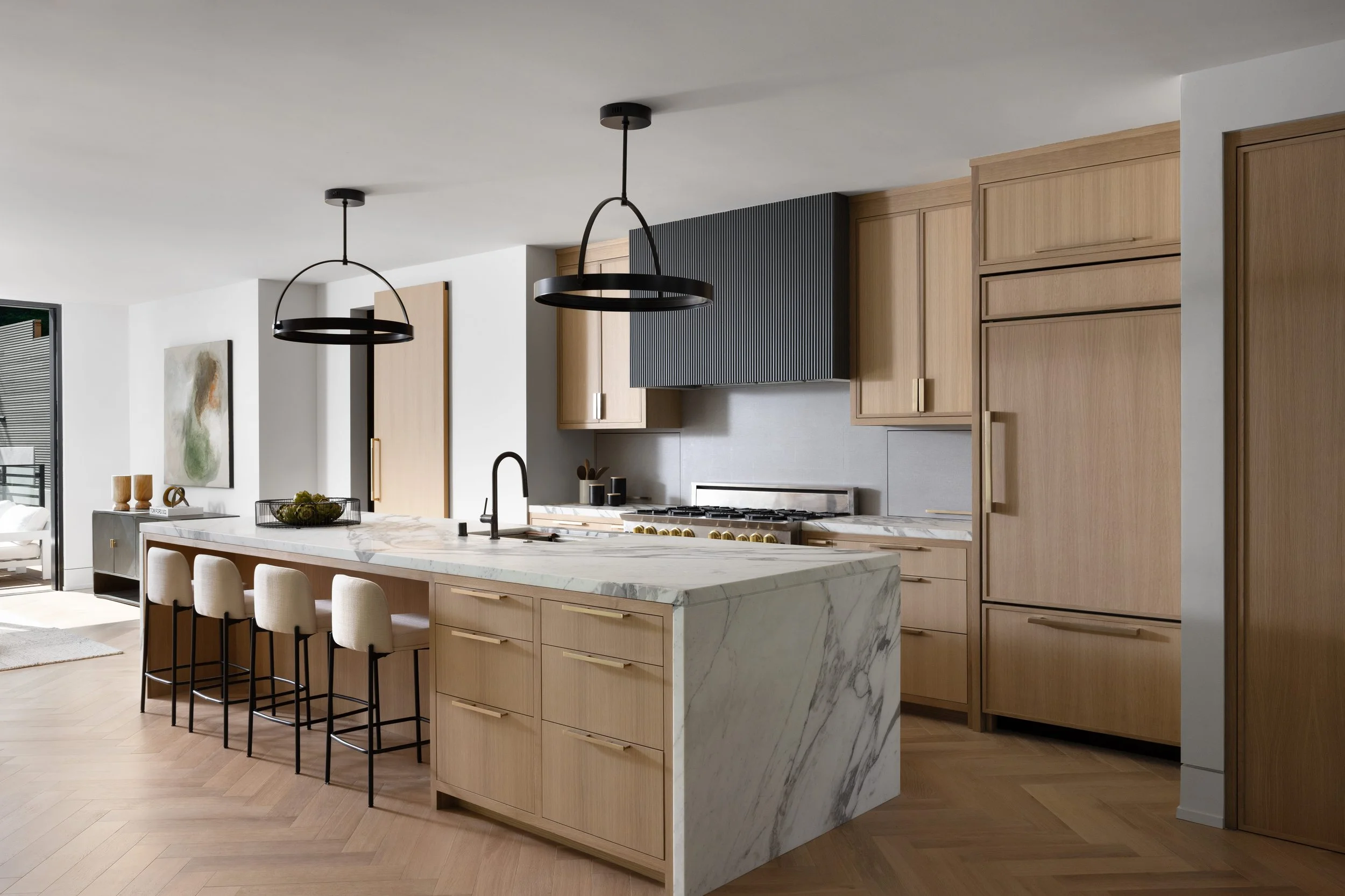 Modern kitchen with wooden cabinets, marble island with seating, black pendant lights, and natural light from a sliding glass door.