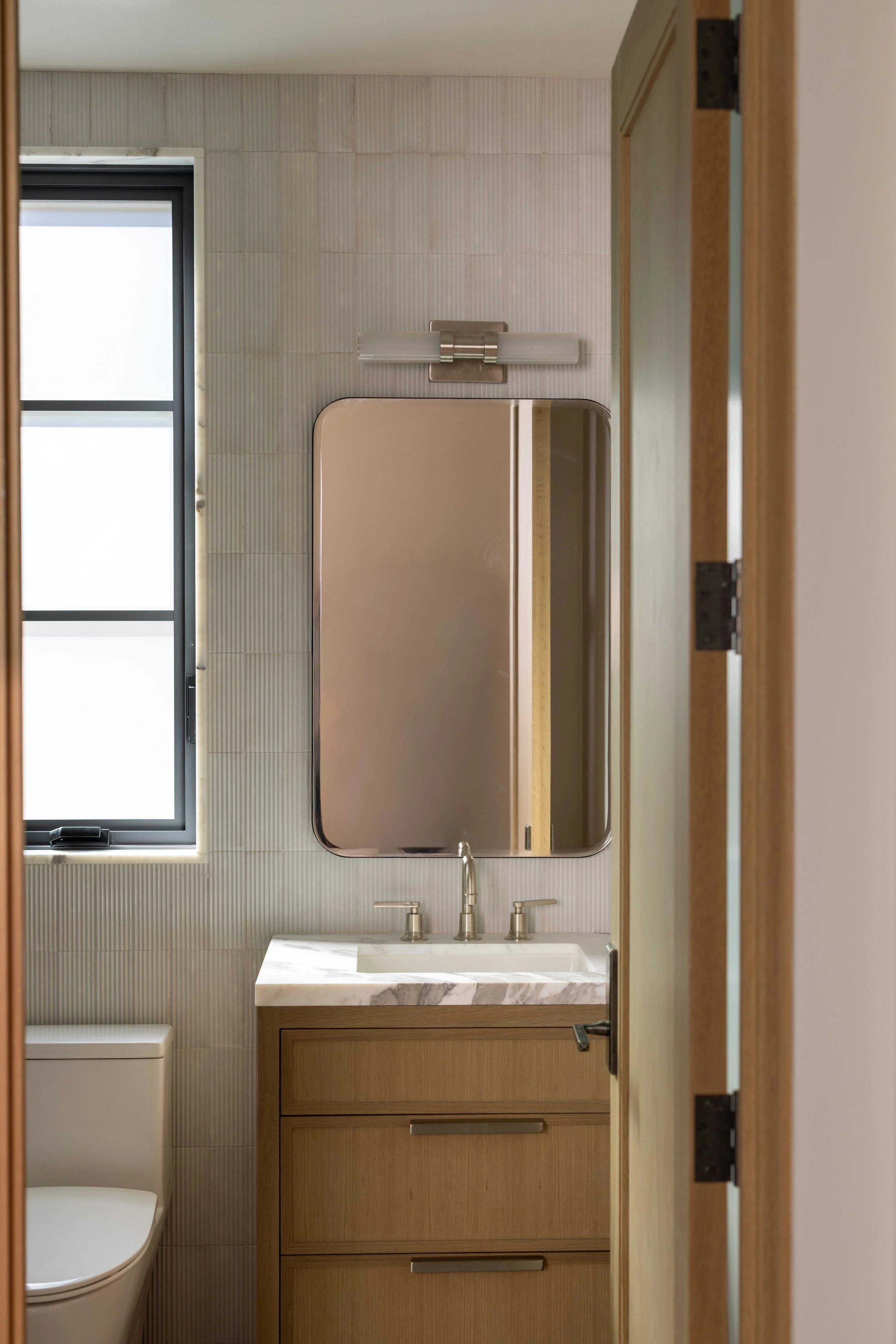 A small bathroom with a wooden vanity, marble countertop, oval mirror, wall-mounted light fixture, window, and a toilet.
