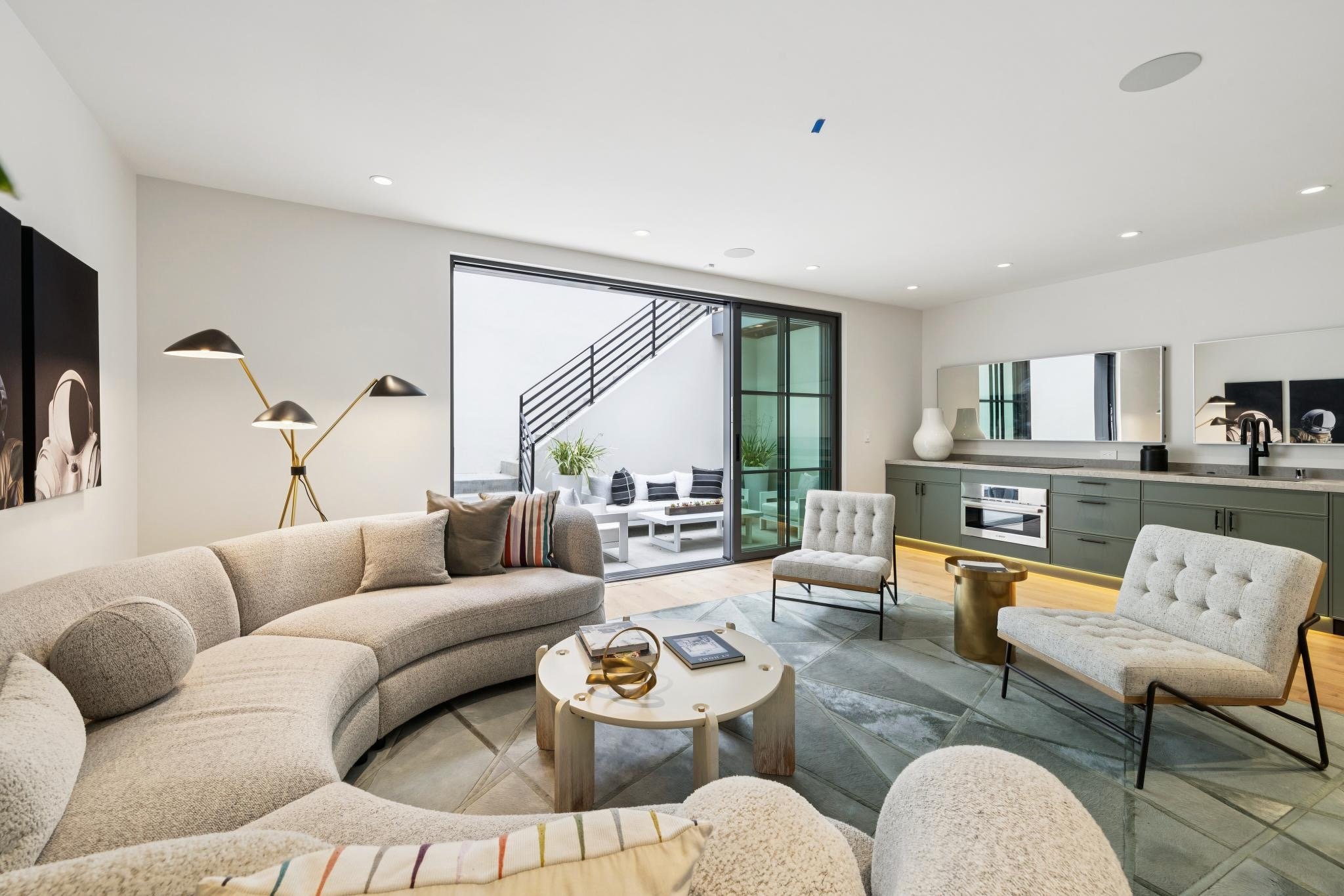 Modern living room with beige sectional sofa, round coffee table, and armchair, open to outdoor patio with seating, sliding glass door, green kitchen cabinets, and decorative vases.