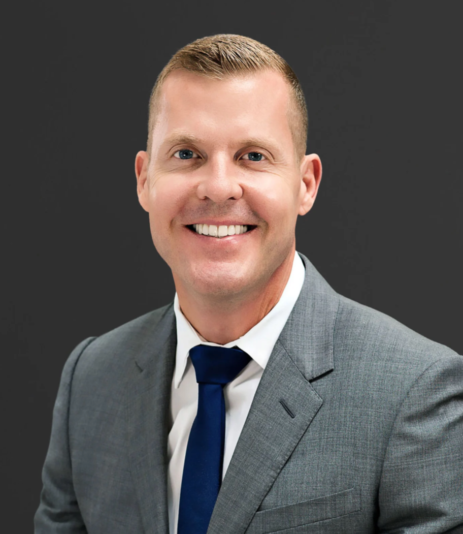A smiling man with short blonde hair dressed in a gray suit, white shirt, and dark blue tie against a dark gray background.