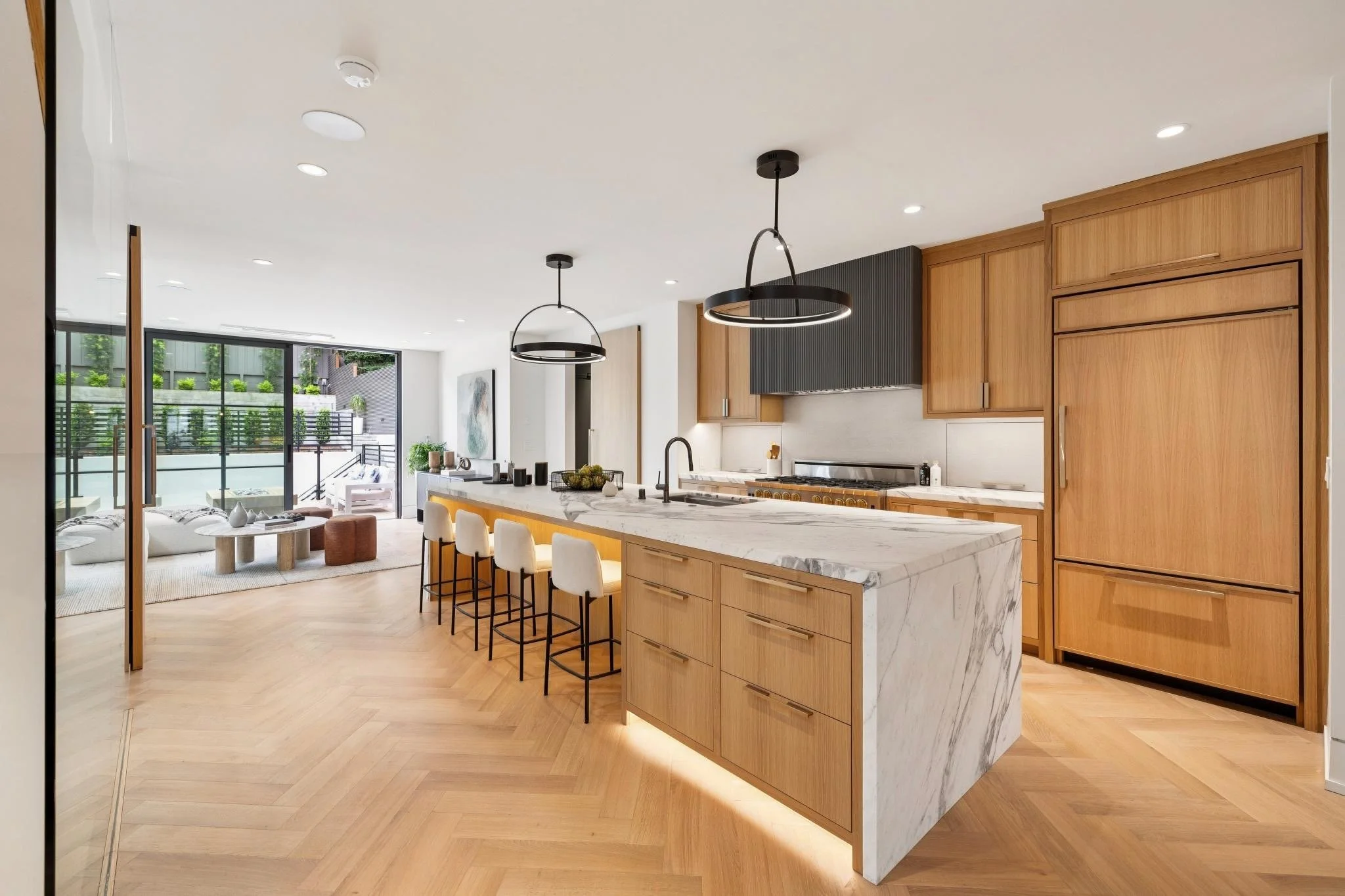 Modern open-concept kitchen and living room with wood cabinetry, a marble kitchen island, pendant lighting, and large sliding glass doors leading to an outdoor patio.