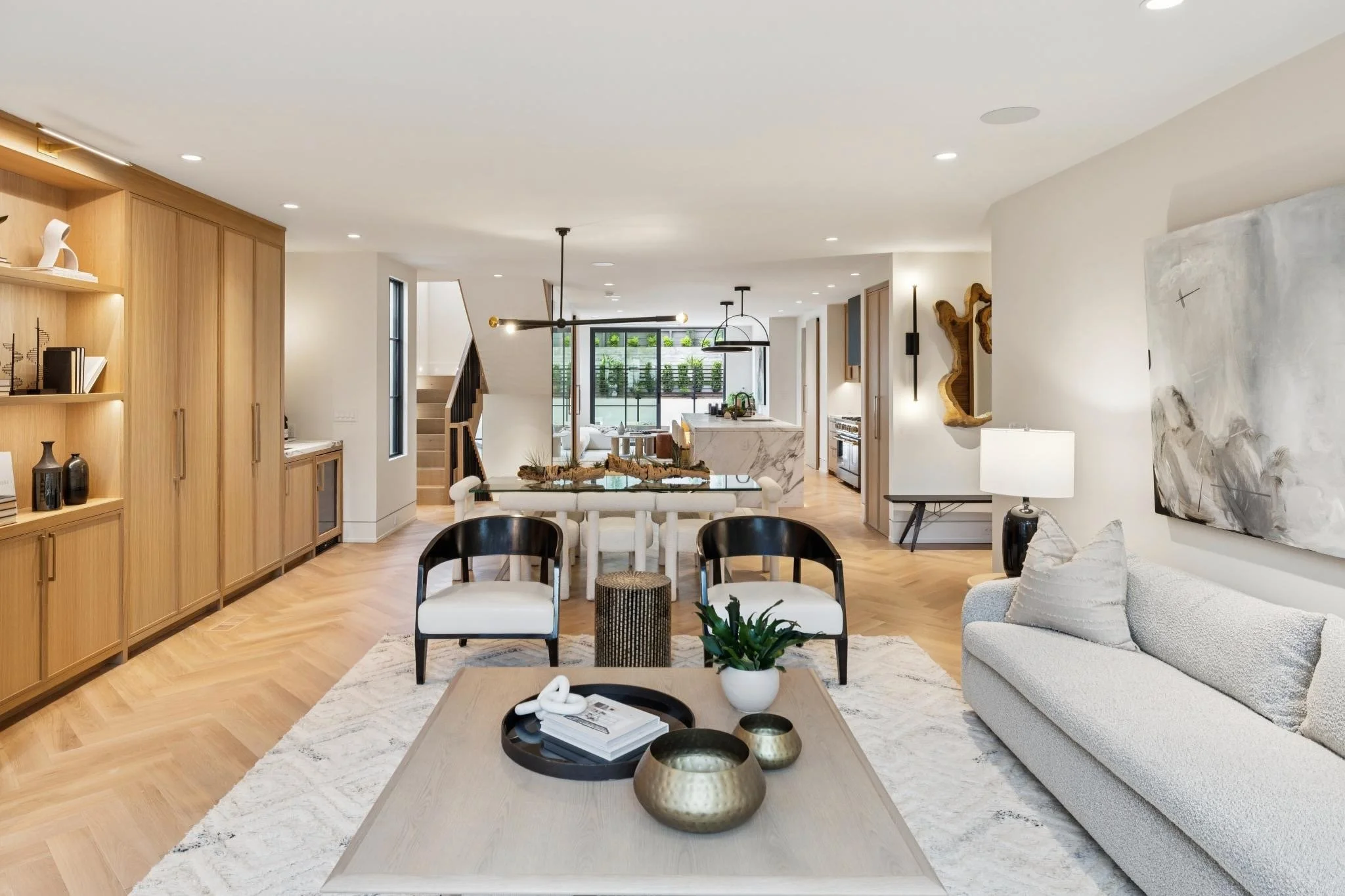 Modern open-concept living room and kitchen with neutral tones, wooden accents, and minimalistic decor, including a white sofa, black and white chairs, abstract artwork, and green plants.