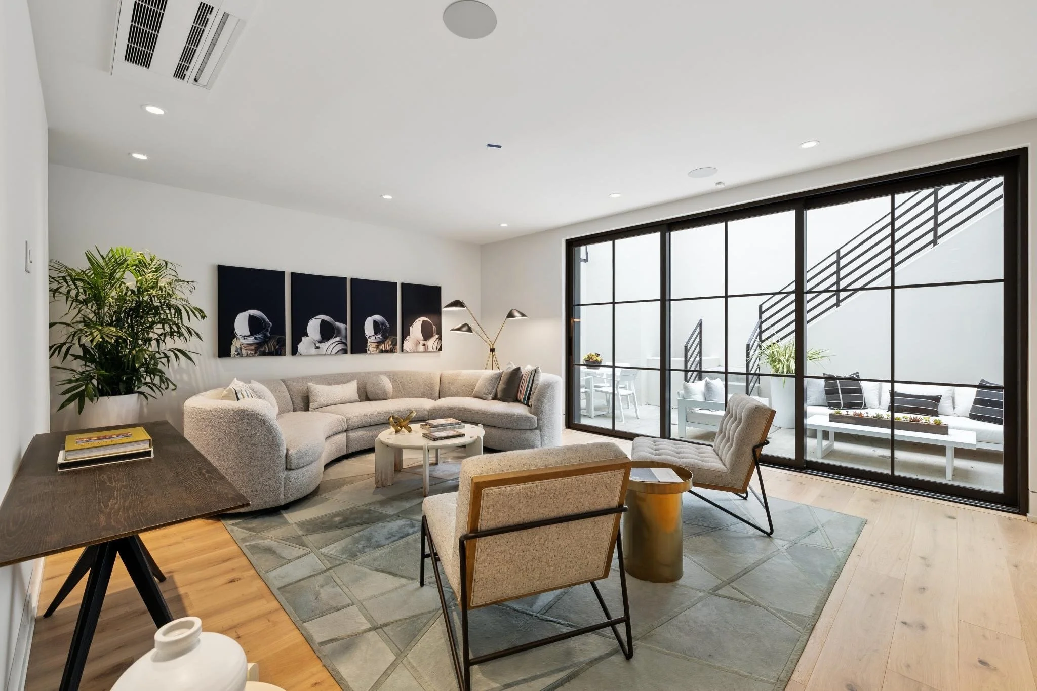 Modern living room with beige sectional sofa, armchair, coffee table, wall art, plants, and large glass doors leading to a balcony with outdoor seating.