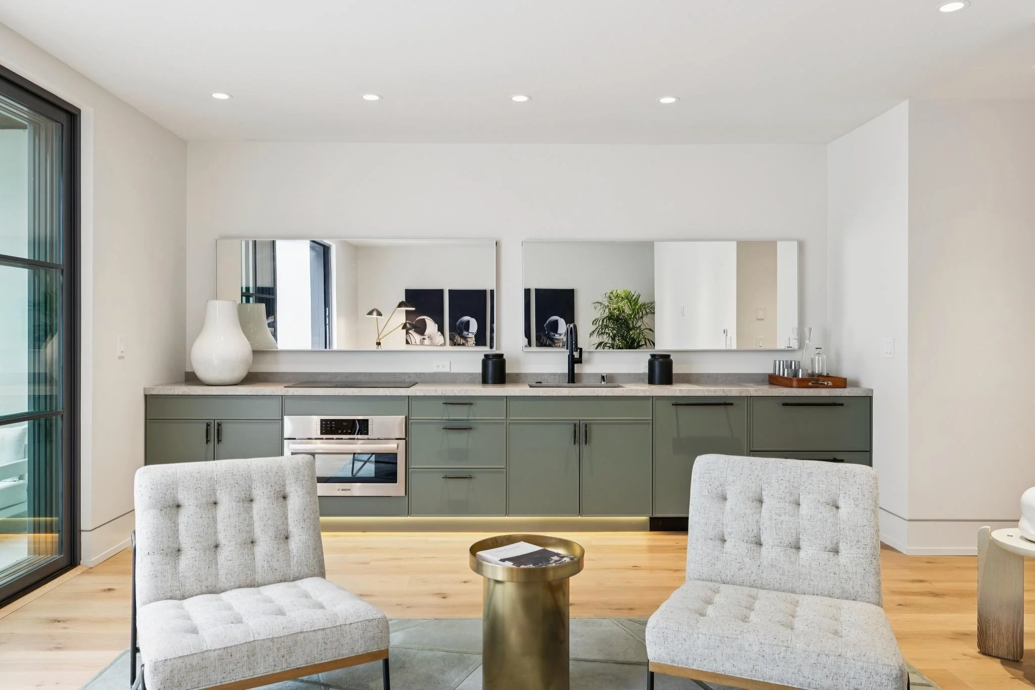 Modern kitchen and dining area with light-colored chairs, a small round gold-colored table, and a kitchen cabinet with a sink, mirrors, and decorative items.