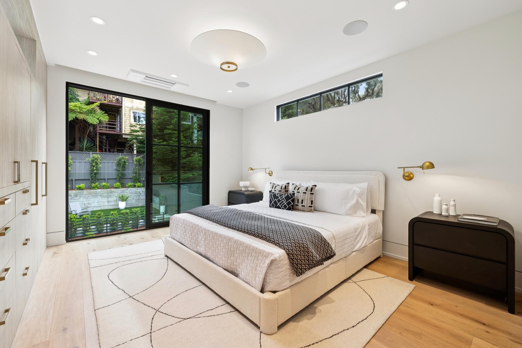 Modern bedroom with a large sliding glass door leading to a balcony, a bed with white linens and decorative pillows, a black nightstand, a white rug with black lines, and minimal wall decor including gold wall sconces.
