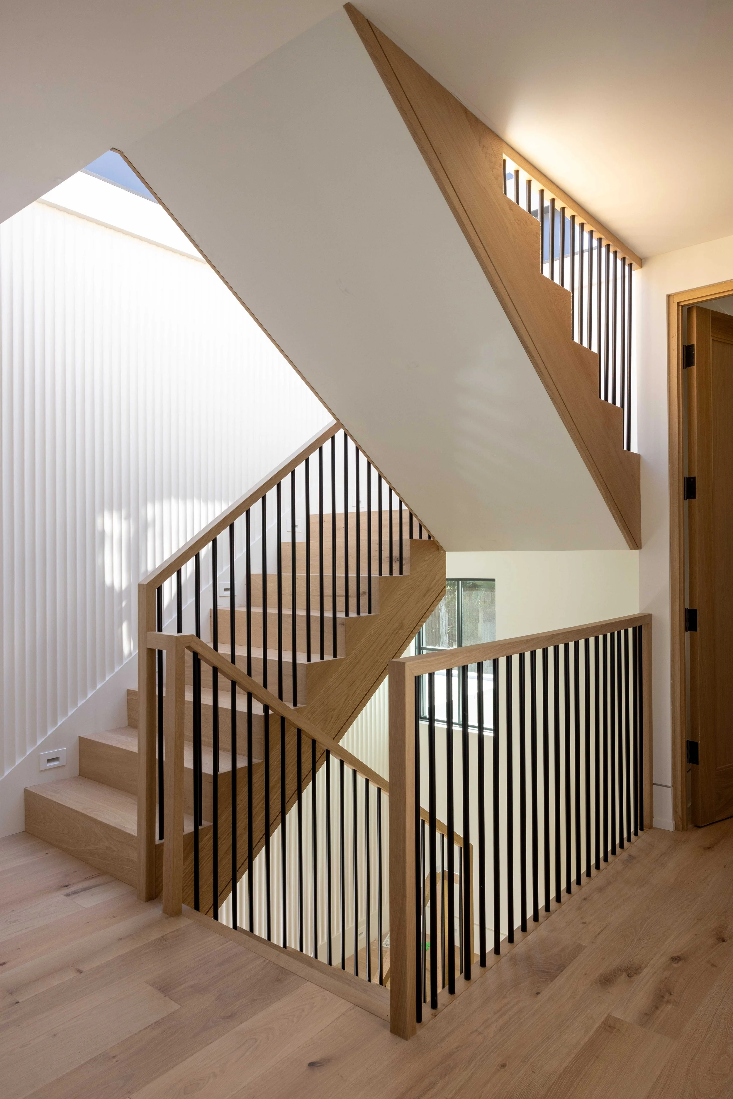 Interior view of a modern staircase with light wood steps, black vertical railings, and a combination of white walls and wooden accents, with a large window natural light.