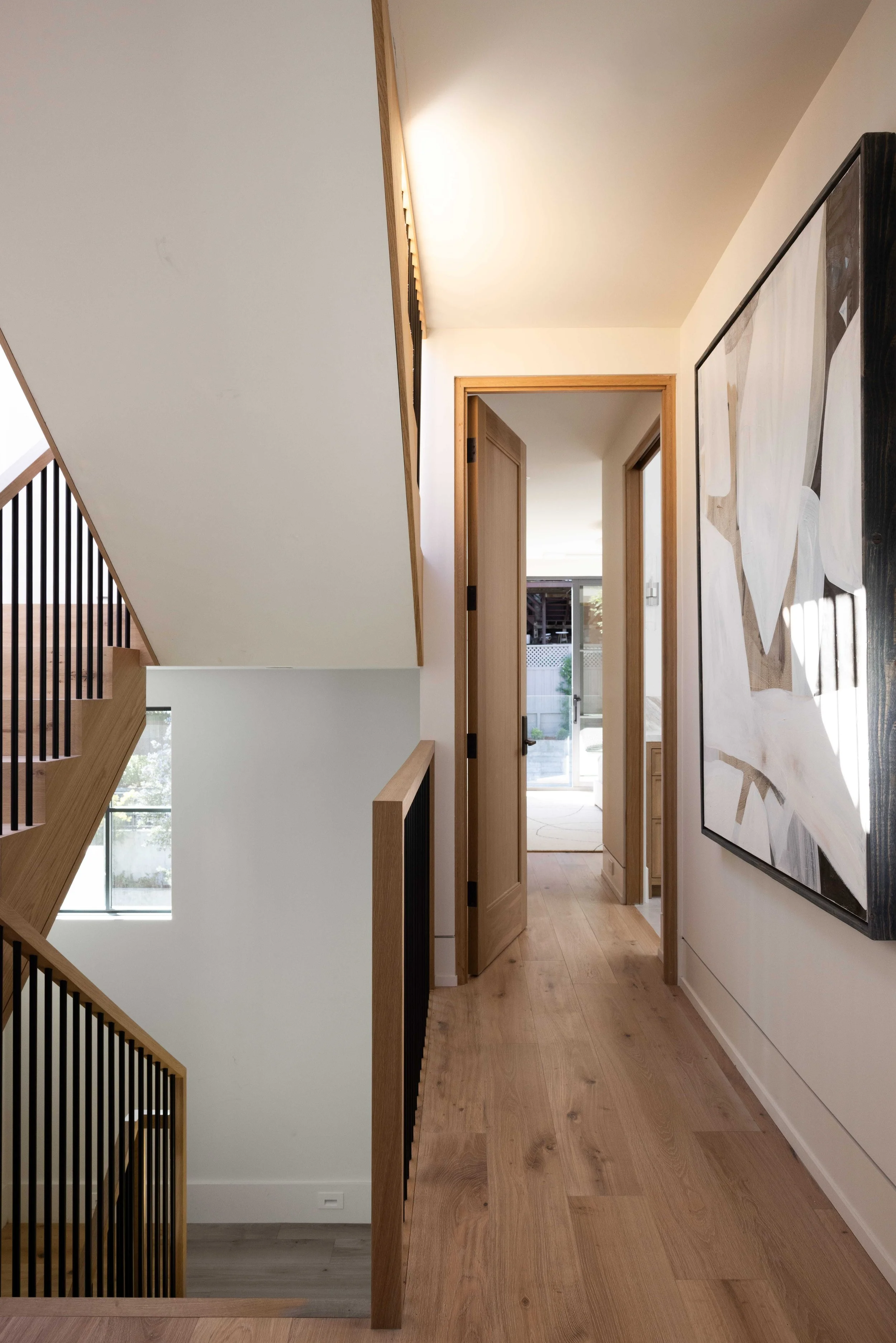 Interior view of a modern house hallway with wooden floors, a door open to a bedroom, and a staircase with wooden railing on the left. A large abstract painting is on the right wall.