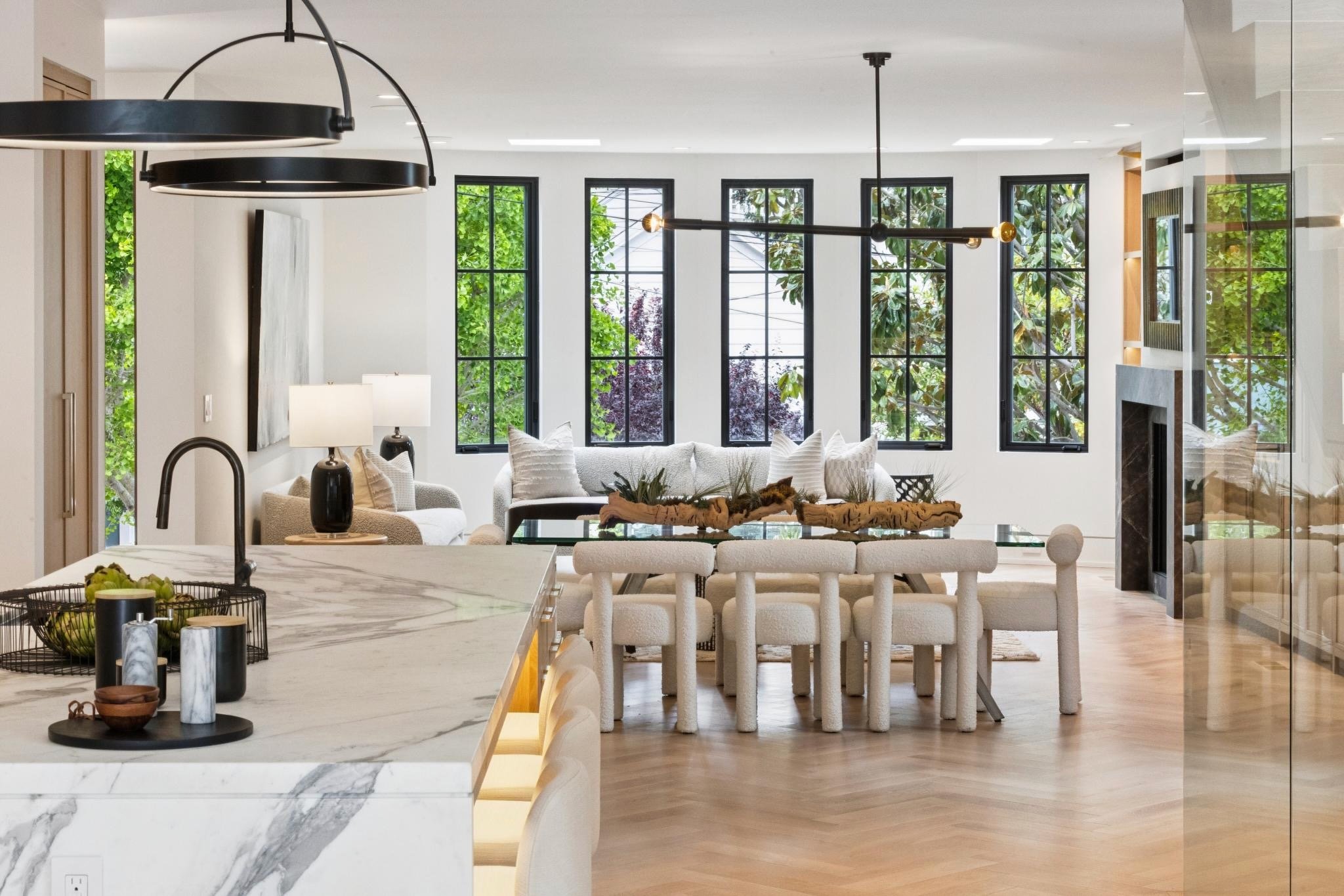 Open-concept living area with white walls, large windows with greenery outside, and modern lighting fixtures.