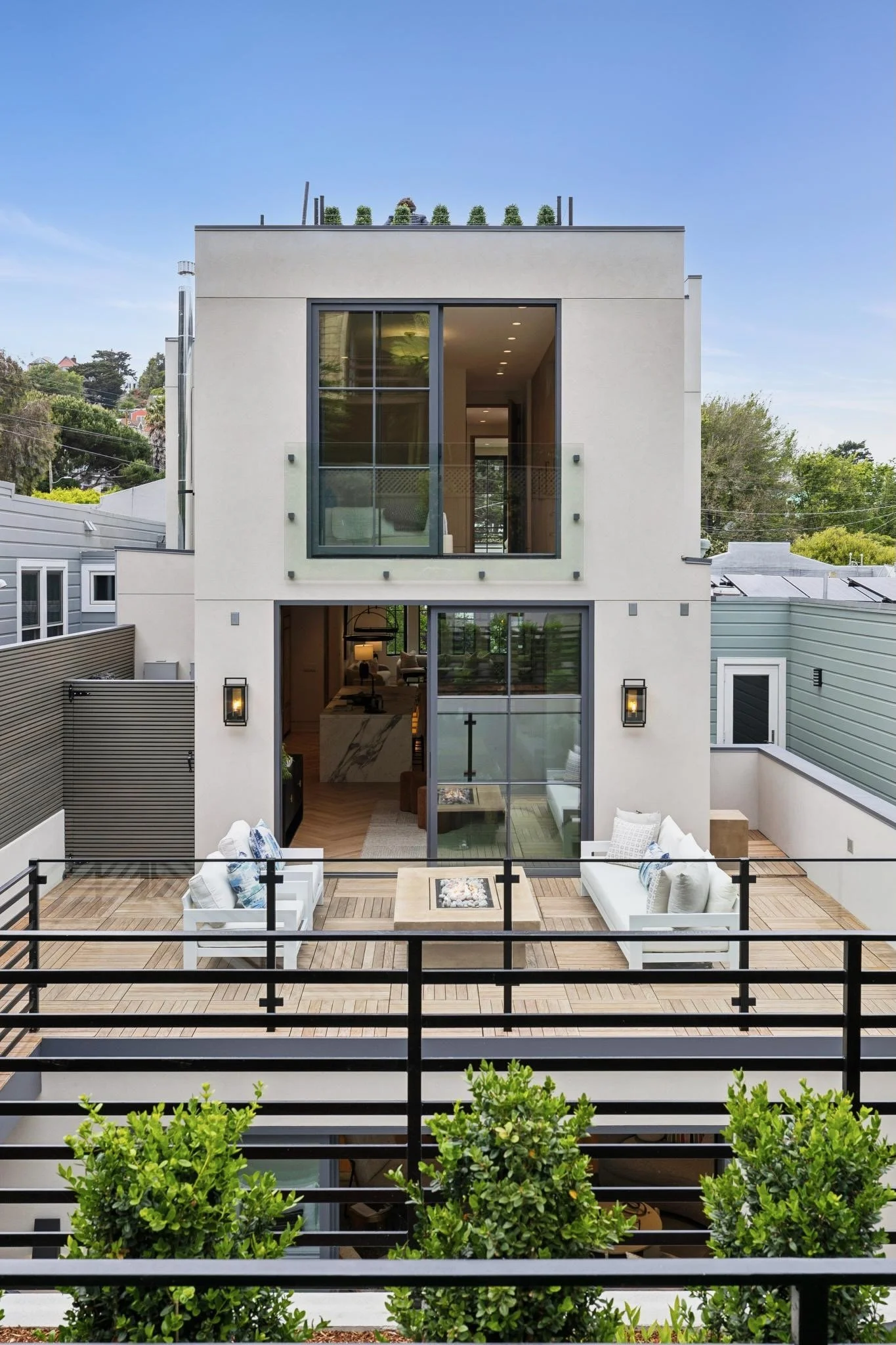 Modern multi-story house with balconies, large windows, outdoor seating area with white furniture, a firepit, and greenery in the foreground.