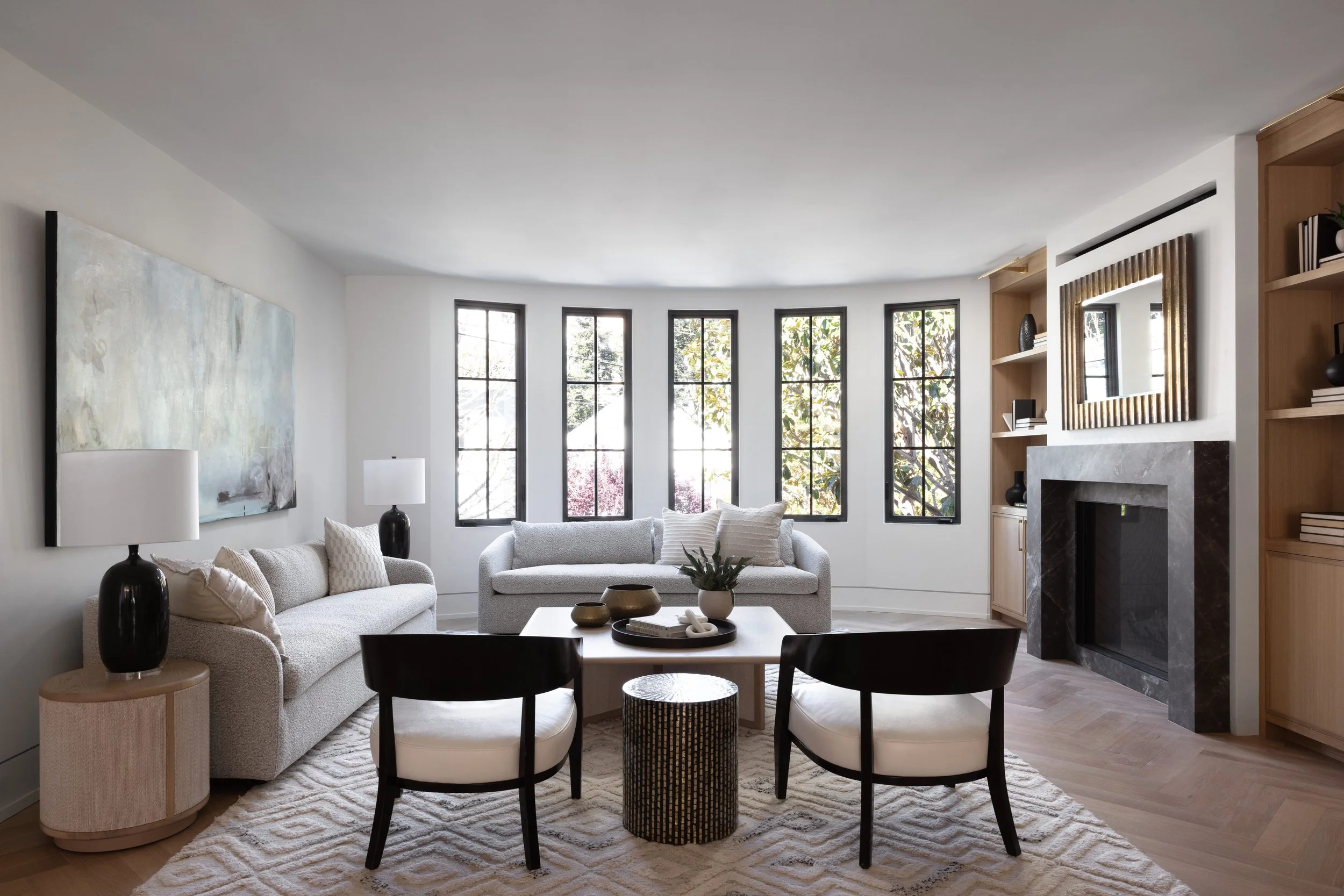 Bright living room with curved wall of large windows, white sofas, black and white chairs, fireplace with black marble surround, wooden shelves, and a large abstract painting on the wall.