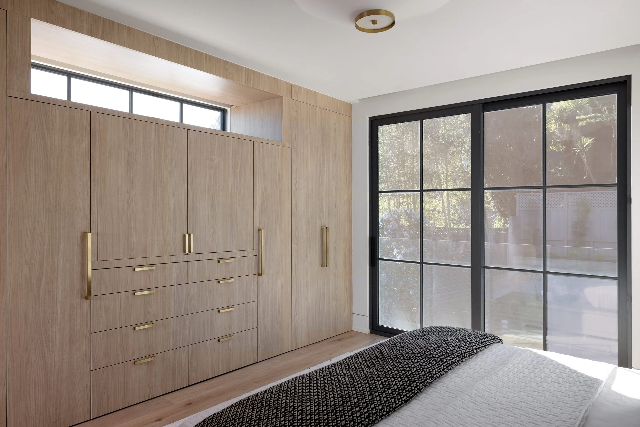 Modern bedroom with light wood built-in wardrobe, large black-framed window, and a bed with white quilt and black patterned blanket.