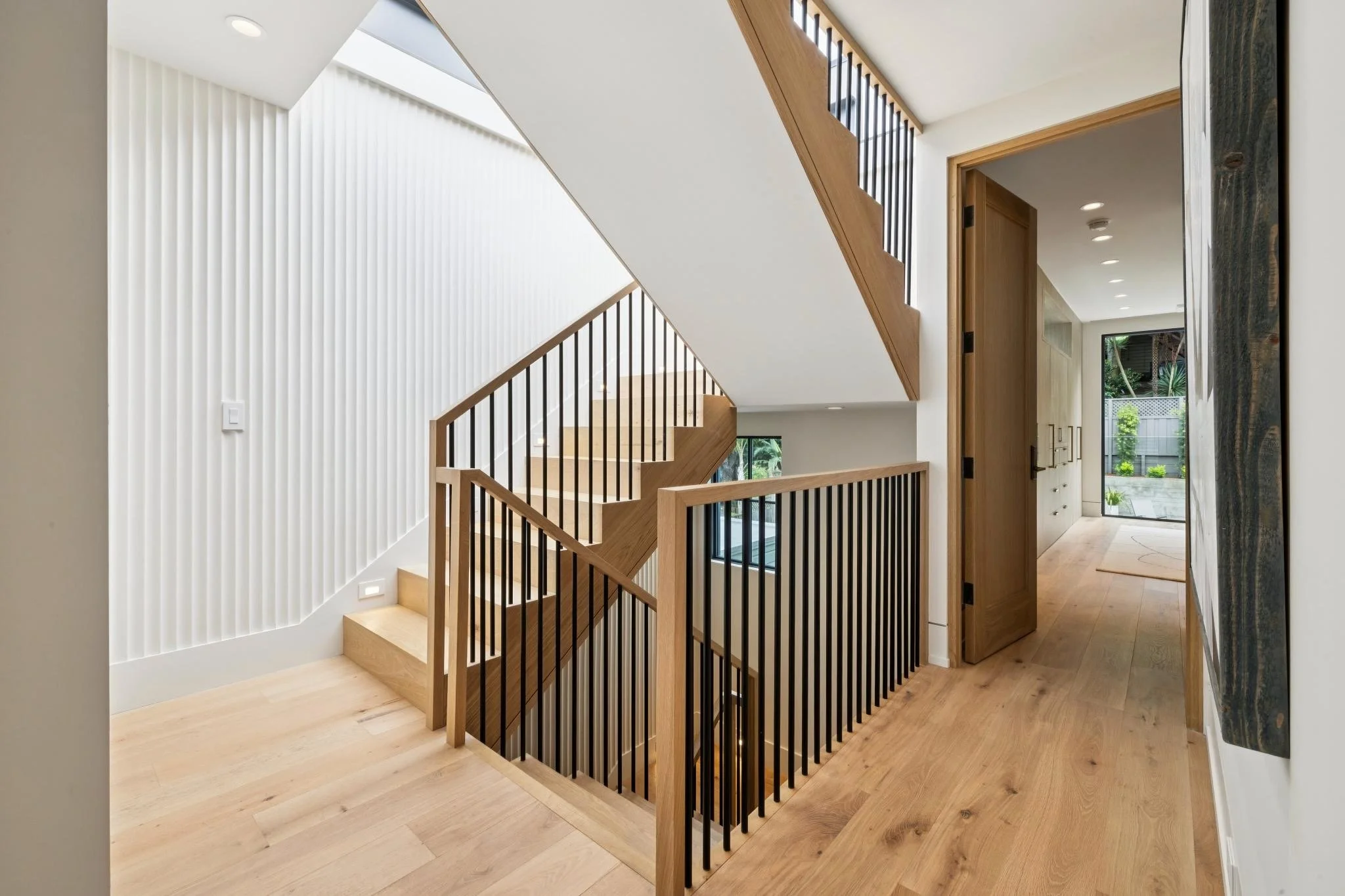 Interior view of a modern home stairway with wooden steps and black metal railing, white walls, natural light from an overhang window, and a door leading to an outdoor space with greenery.