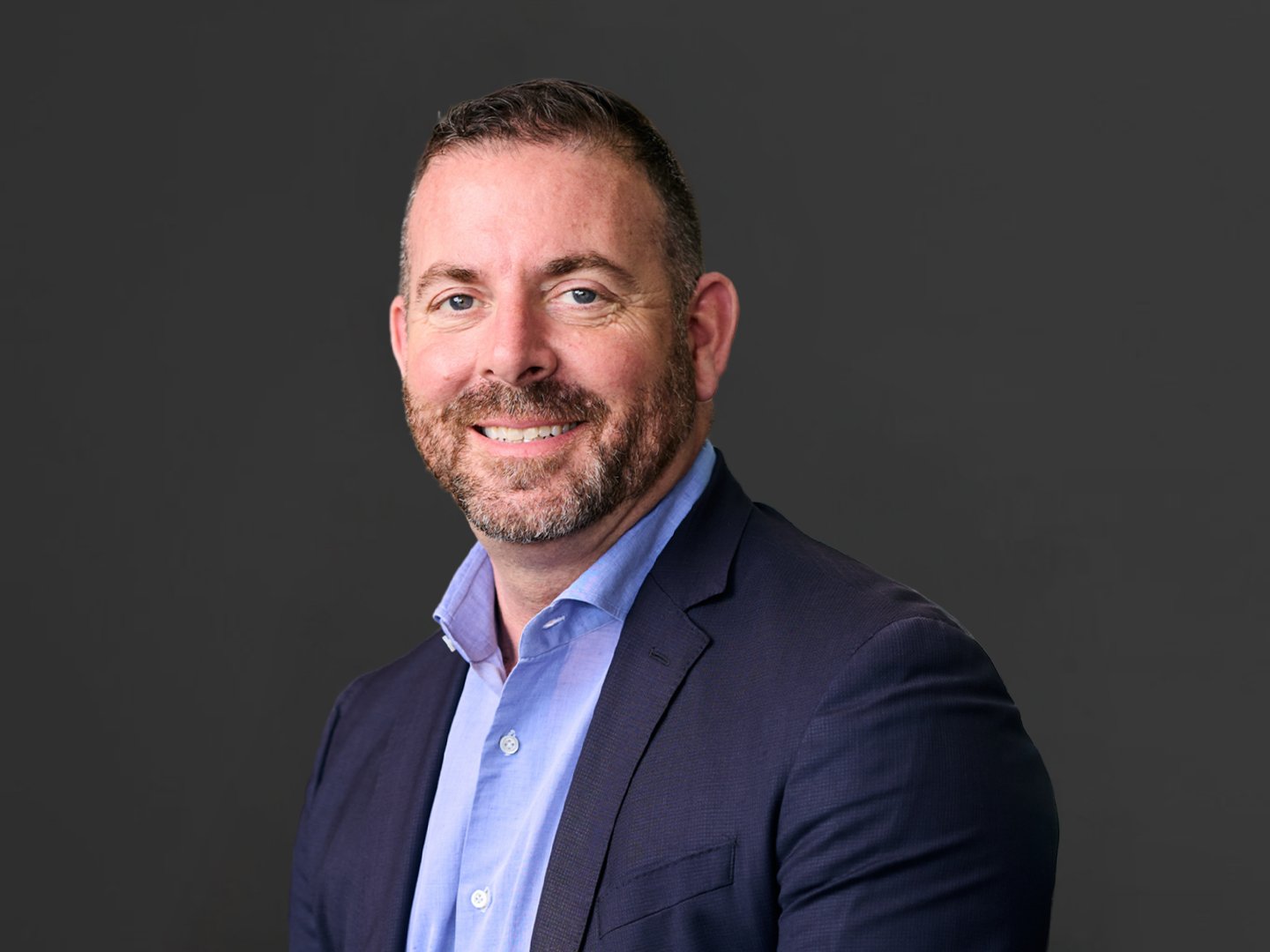 Headshot of a smiling man with short brown hair, a beard, wearing a navy blazer and light blue shirt, against a dark gray background.