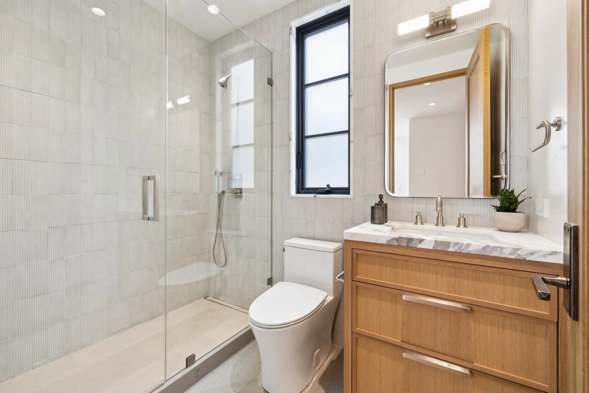 Modern bathroom with glass-enclosed shower, white tiled walls, a window, a wooden vanity with a marble countertop, a mirror, a plants, and a toilet.