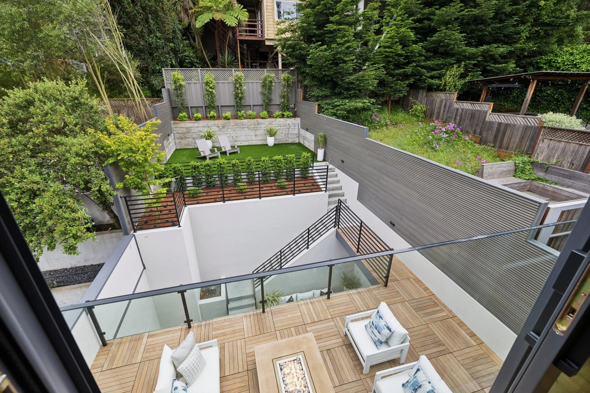 View of a modern multi-tiered backyard patio with decking, outdoor chairs, and a small table, surrounded by lush green trees and a wooden fence.