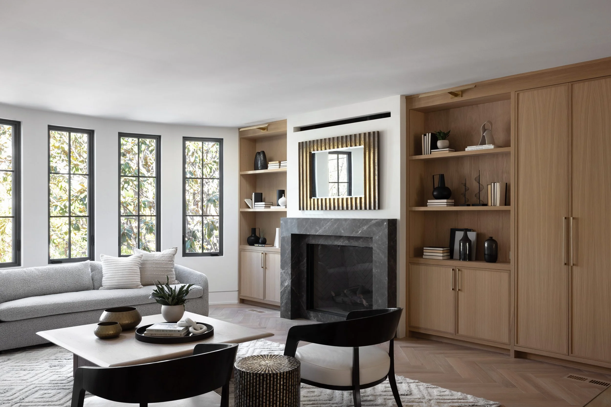 Living room with large windows, a grey sofa with white pillows, a wood coffee table with decorative bowls and a plant, a black and white chair, built-in wooden shelves with decorative items, and a fireplace with a mirror above it.