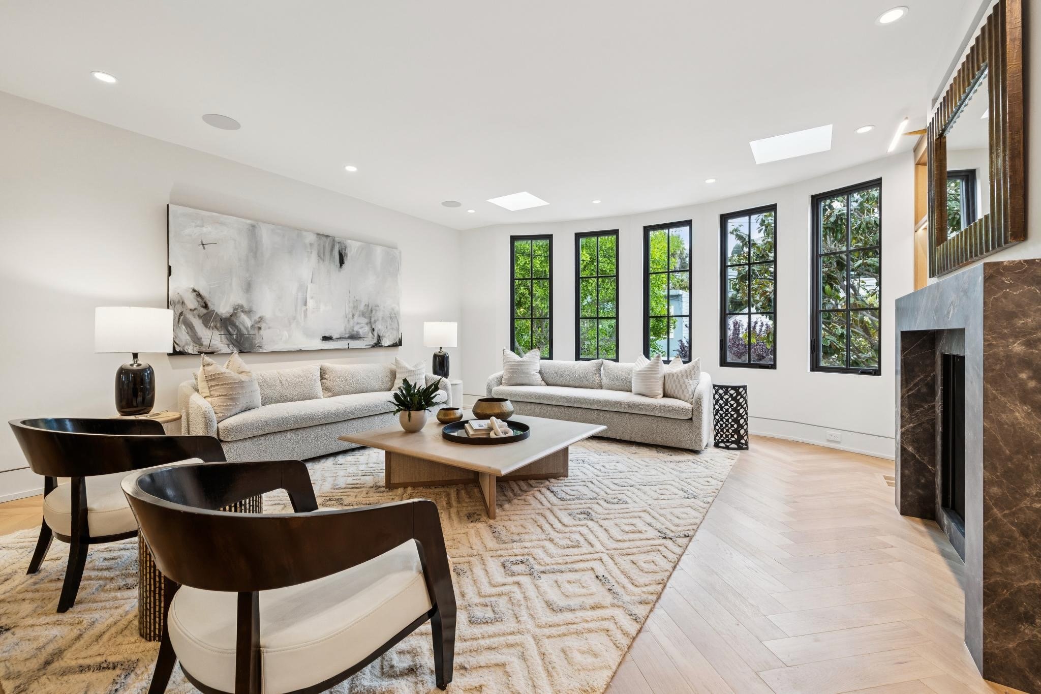 Living room with white sofas, black armchairs, a low coffee table, two table lamps, modern artwork, large windows, and a stone fireplace.