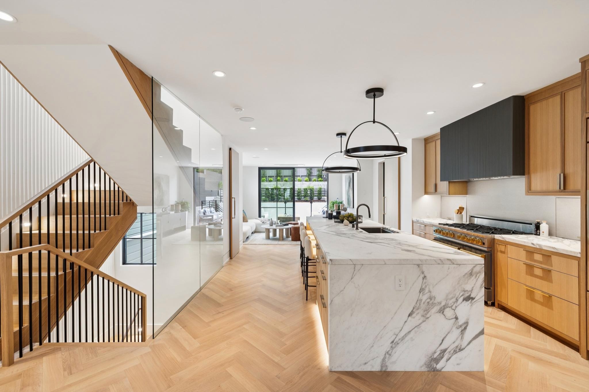 Modern open-concept kitchen with marble island, wooden cabinets, black hanging lights, and views of a living room and backyard outside large windows.