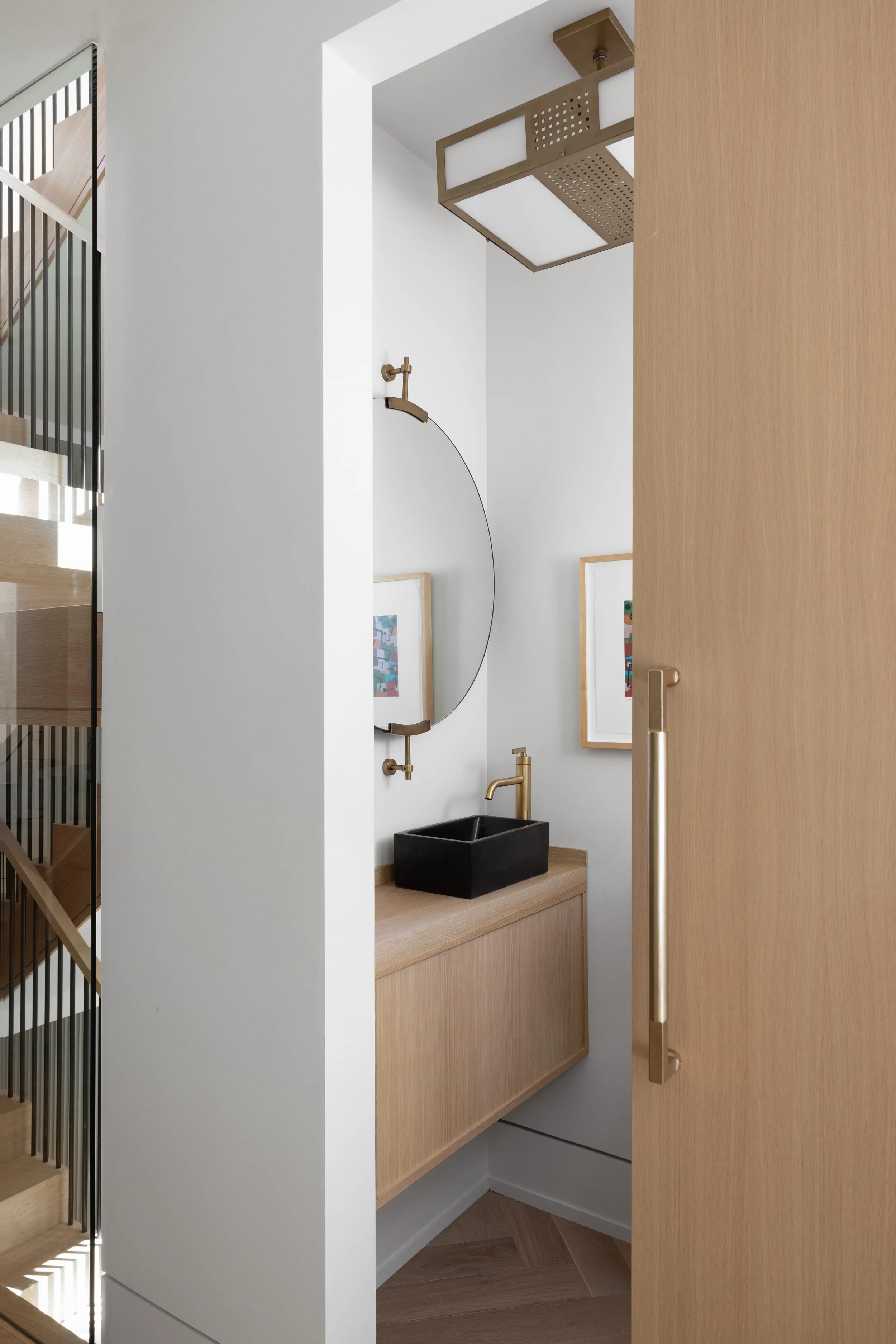 A small bathroom with a black vessel sink, gold faucet, round mirror, and two framed artworks on the wall. The wooden vanity and door have a light wood finish, and there's a modern ceiling light fixture.