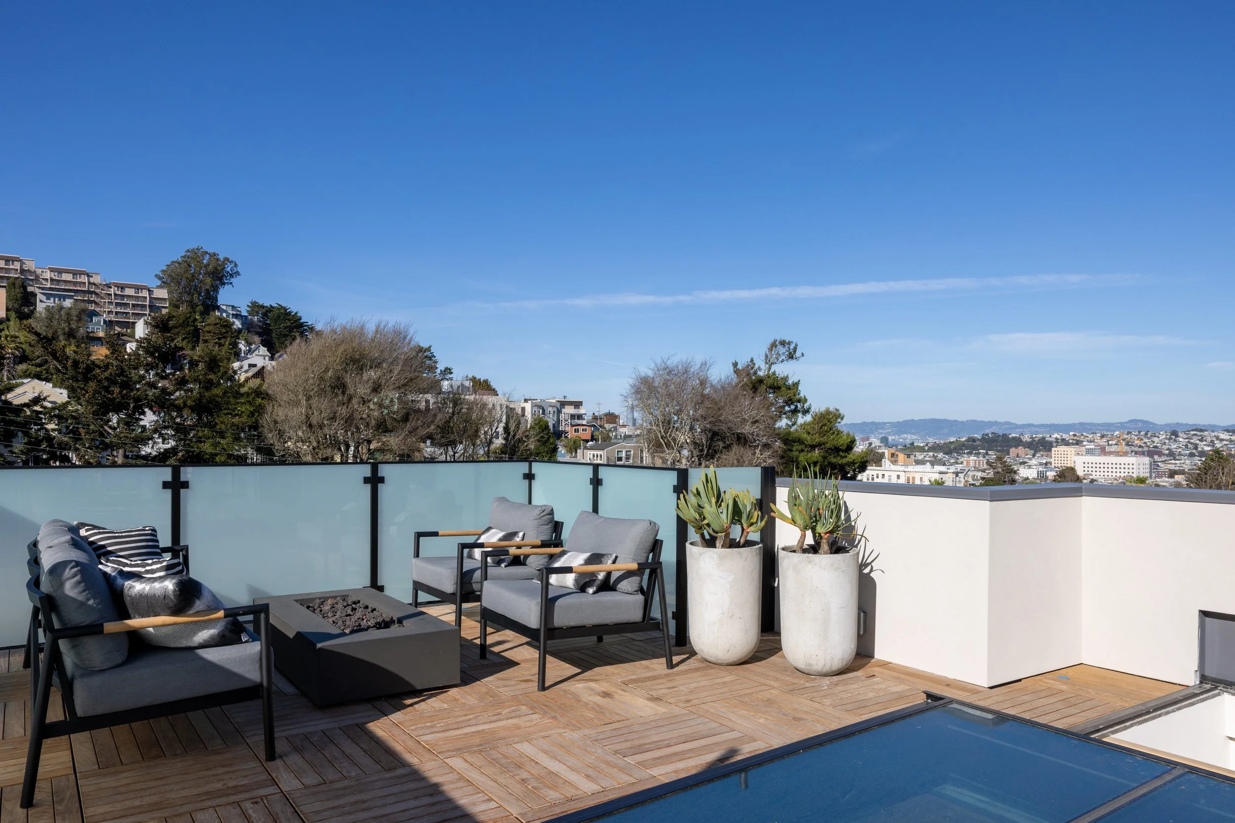 Rooftop patio with modern outdoor furniture, including gray chairs and a fire pit, decorated with pillows and two large potted plants, overlooking a cityscape with trees and buildings under a blue sky.