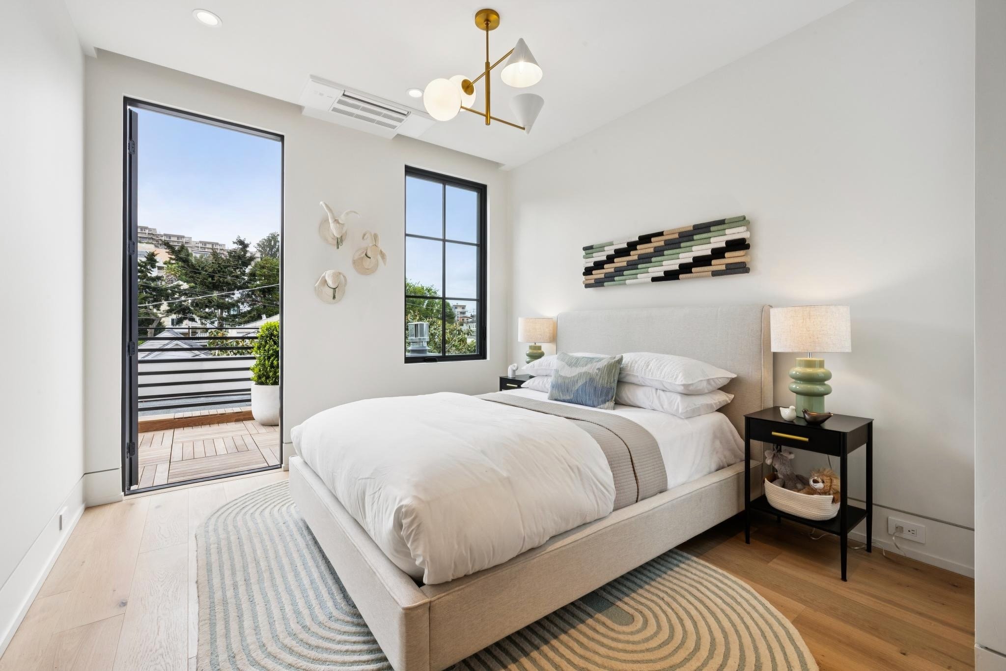 A modern bedroom with a large white bed, black side table, green and white ceramic table lamps, wall art above the bed, and a balcony door showing an outdoor view with trees and apartment buildings.