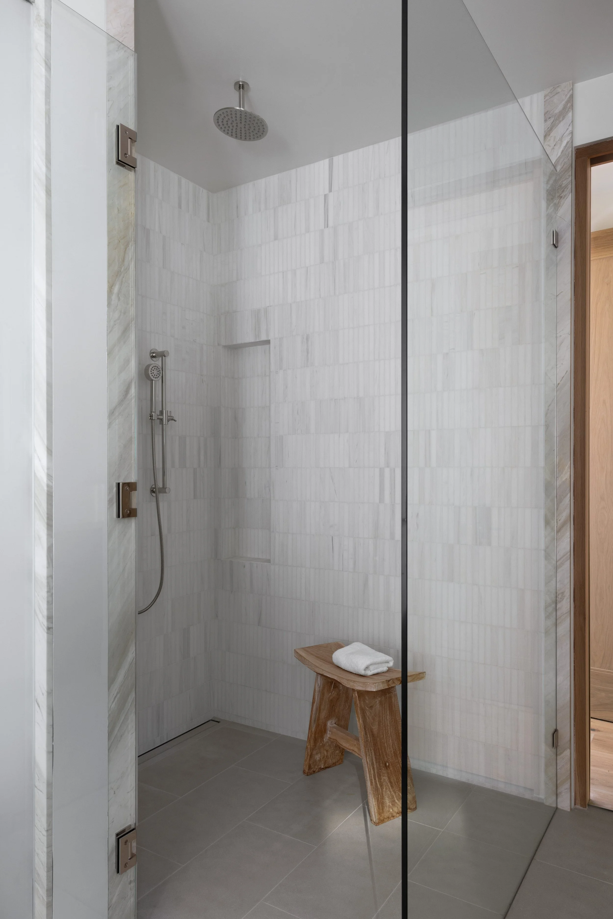 Modern shower with a rainfall showerhead, handheld shower, and tiled walls. A small wooden bench with a towel on it is inside the shower area.