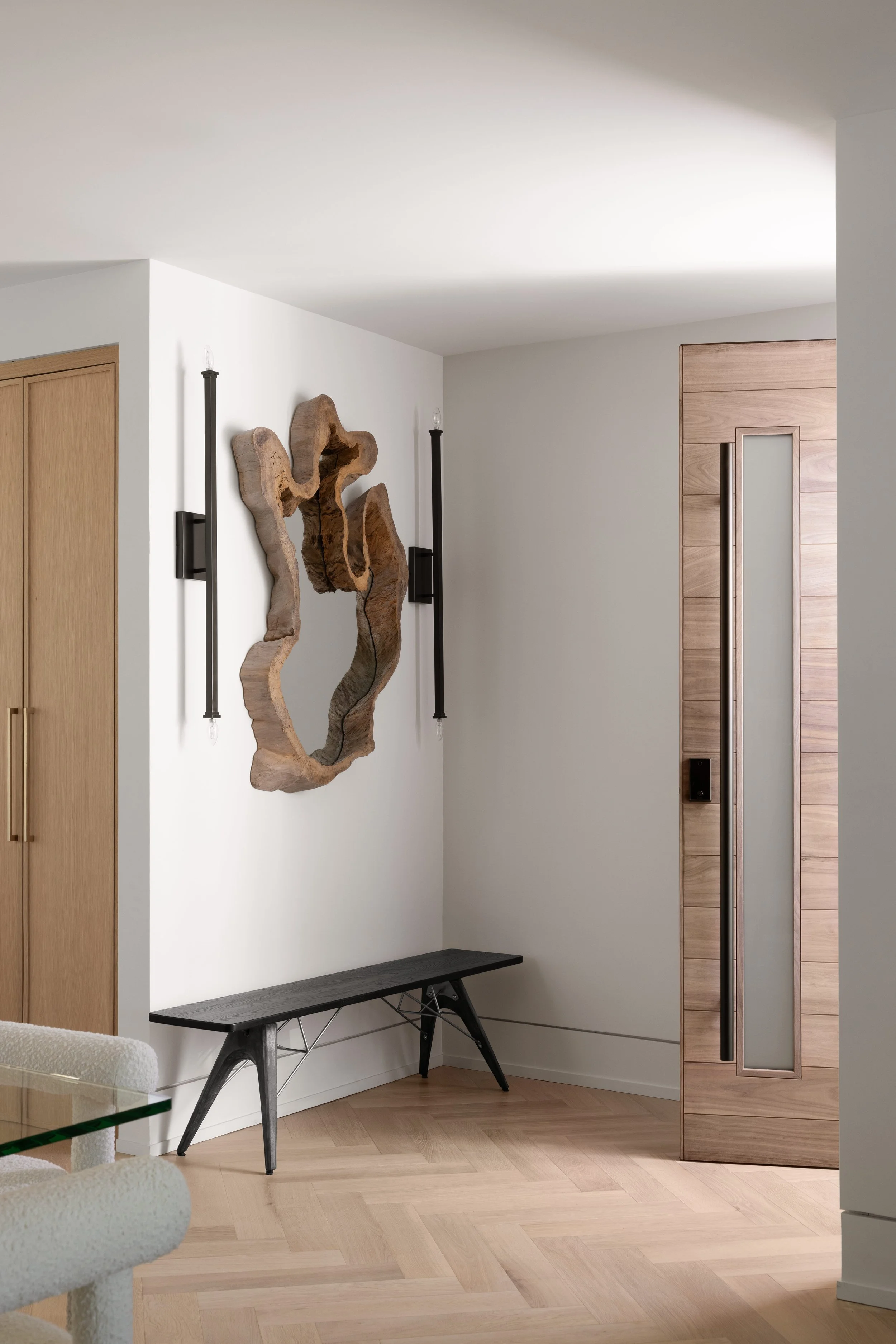 Modern interior entryway with wooden flooring, white walls, a black bench, wood wall art, wall-mounted black light fixtures, and a wooden door with a vertical frosted glass window.