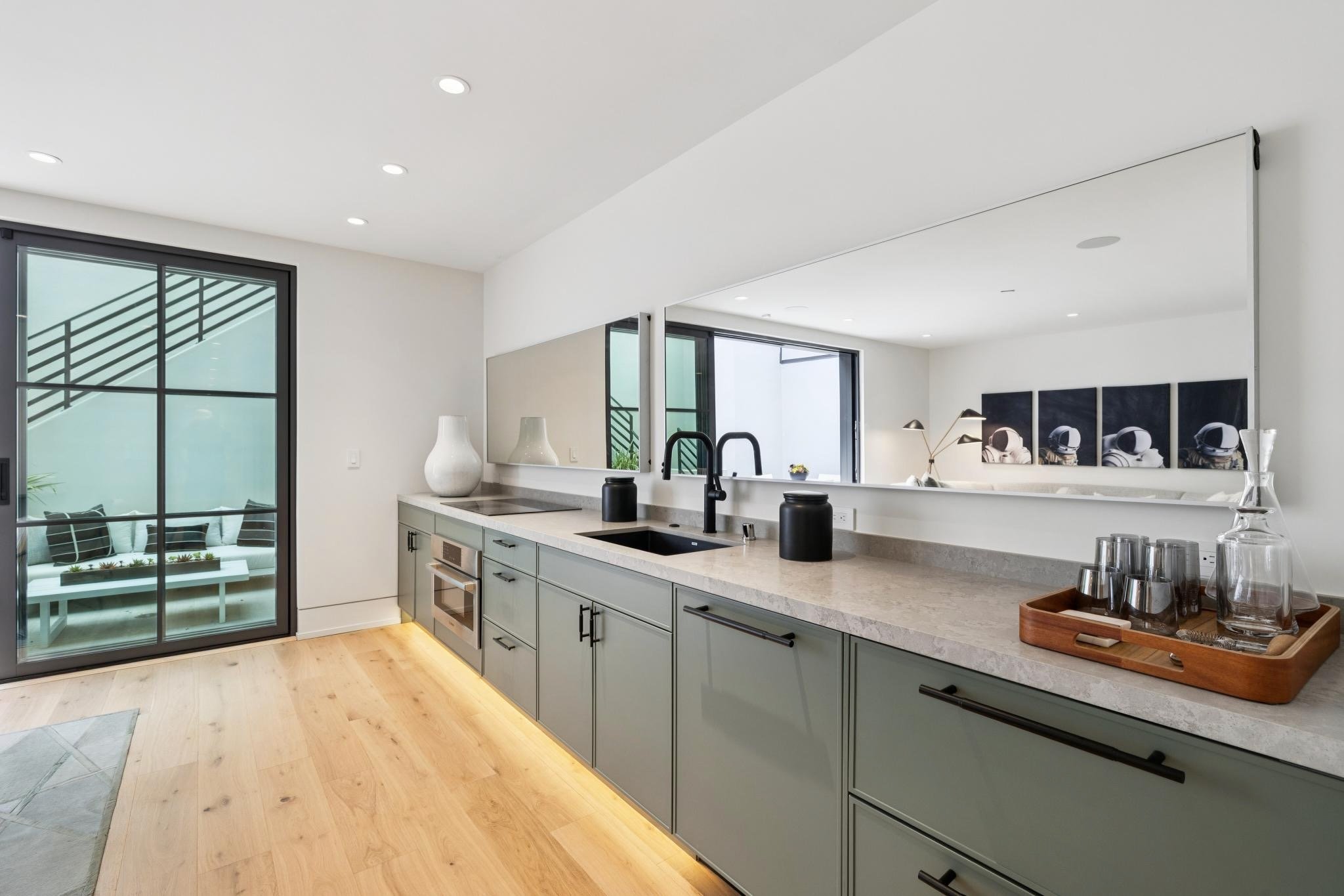 Modern kitchen with light green cabinetry, a marble countertop, black faucet, and a large opening to a bright living area, with framed astronaut artwork on the wall.