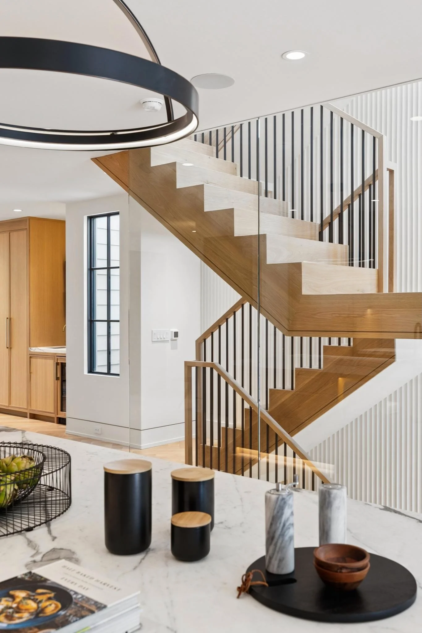 Modern interior with a staircase featuring wooden steps and black metal railings, white walls, and sleek lighting fixtures. There is a kitchen island with decorative black and marble accessories, and a window with black framing.