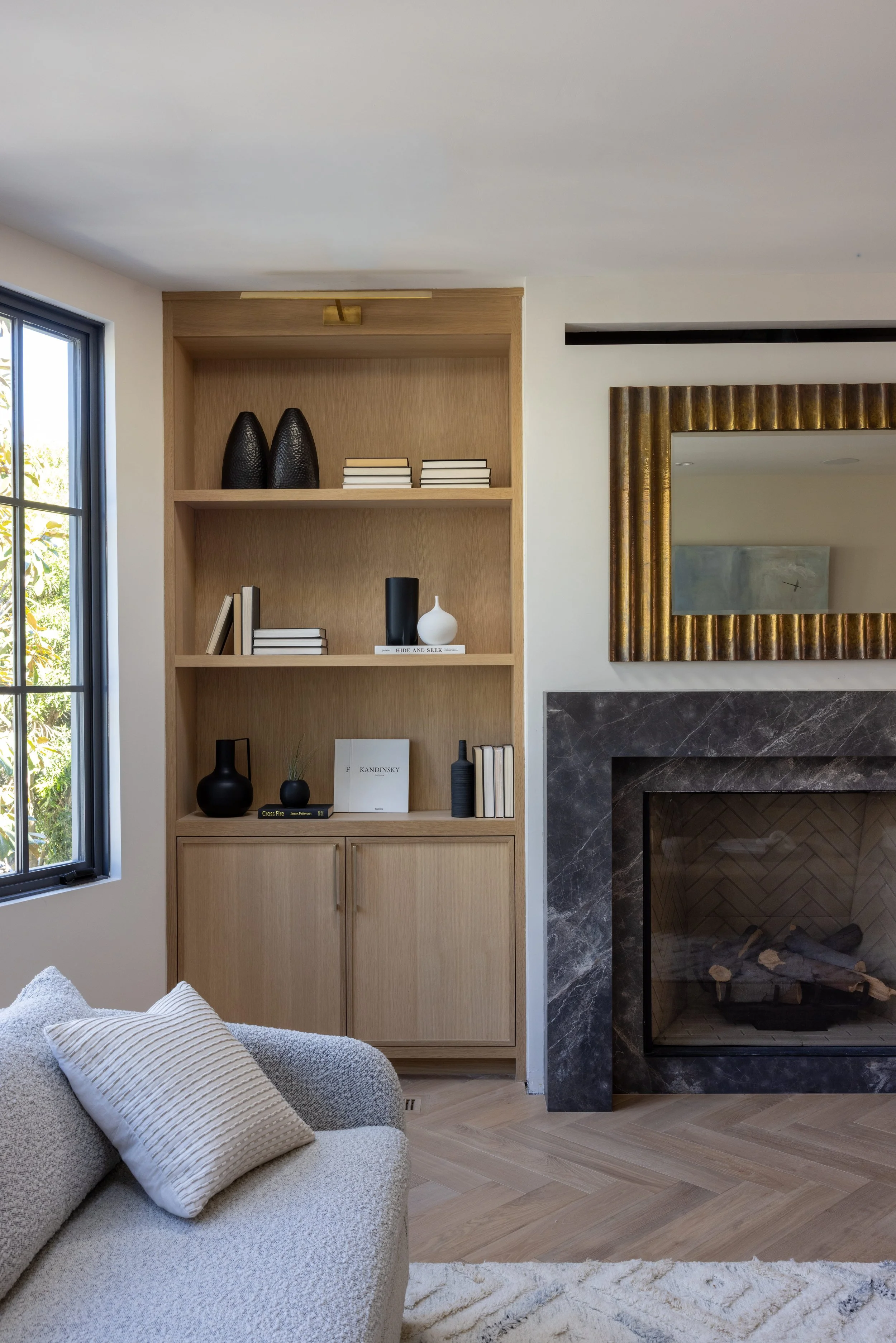 Living room with a beige bookcase, black and white decorative objects, books, a fireplace with dark marble surround, large mirror with gold frame, window on the left, and a white couch with a striped pillow.
