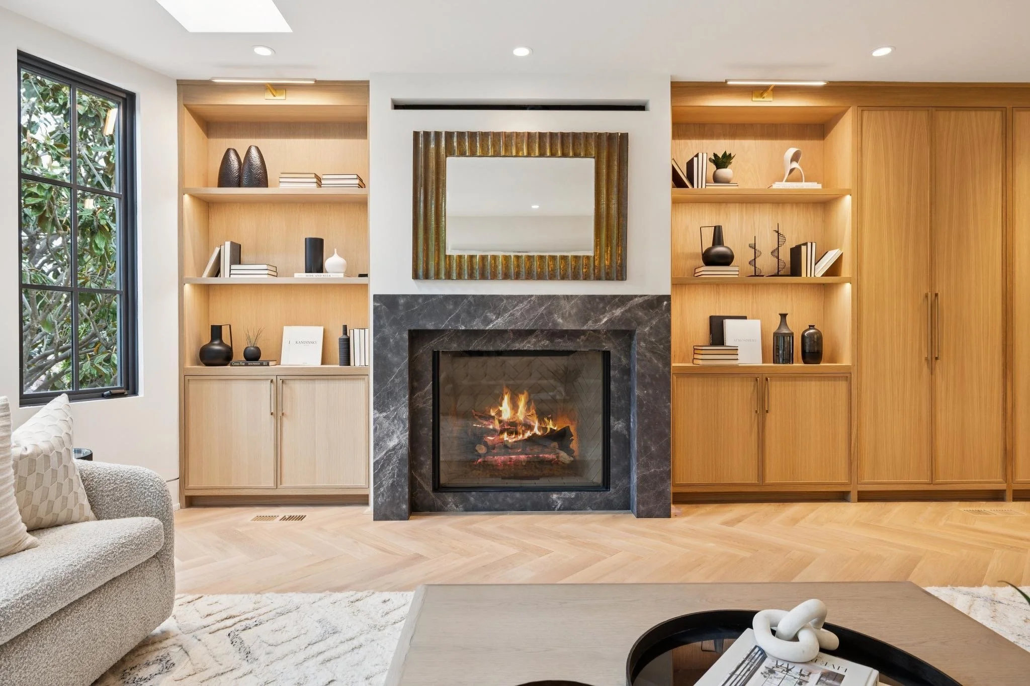 Living room with a fireplace, built-in wooden shelves with decor and books, a window with greenery outside, a beige sofa, and a light-colored wooden floor.