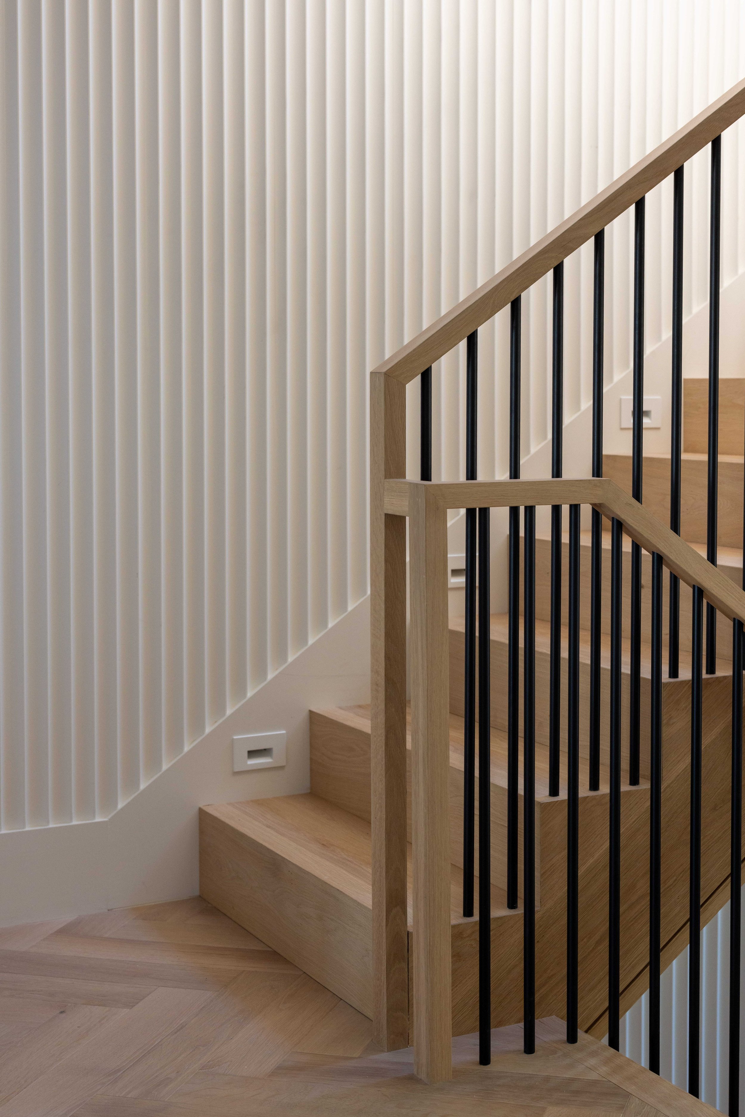 Interior staircase with light wood steps, black metal balusters, and a light wood handrail. The wall beside the stairs has vertical white slats, and there is a white electrical outlet near the bottom of the stairs.