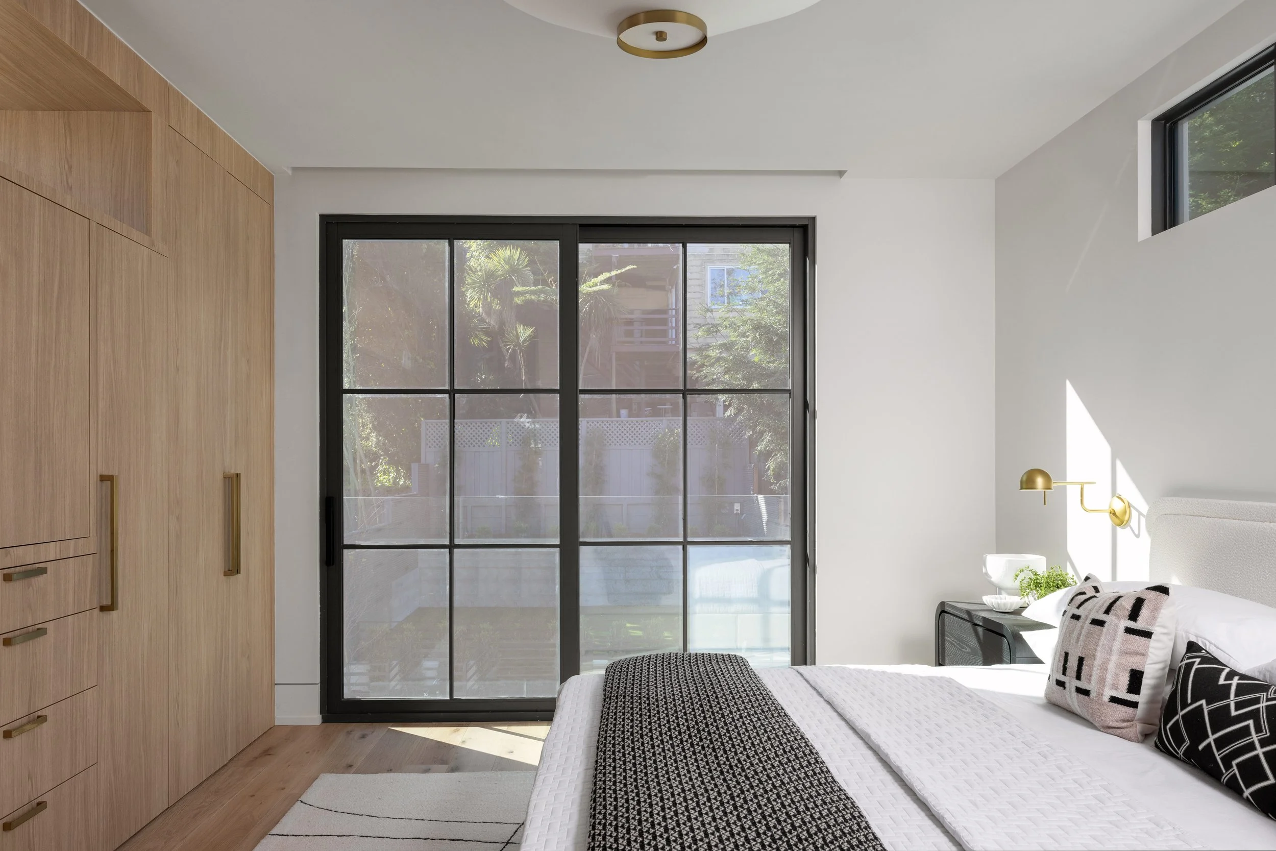 Minimalist bedroom with a large sliding glass door, wooden built-in wardrobe, bed with black and white pillows, bedside table, and gold wall-mounted lamp.