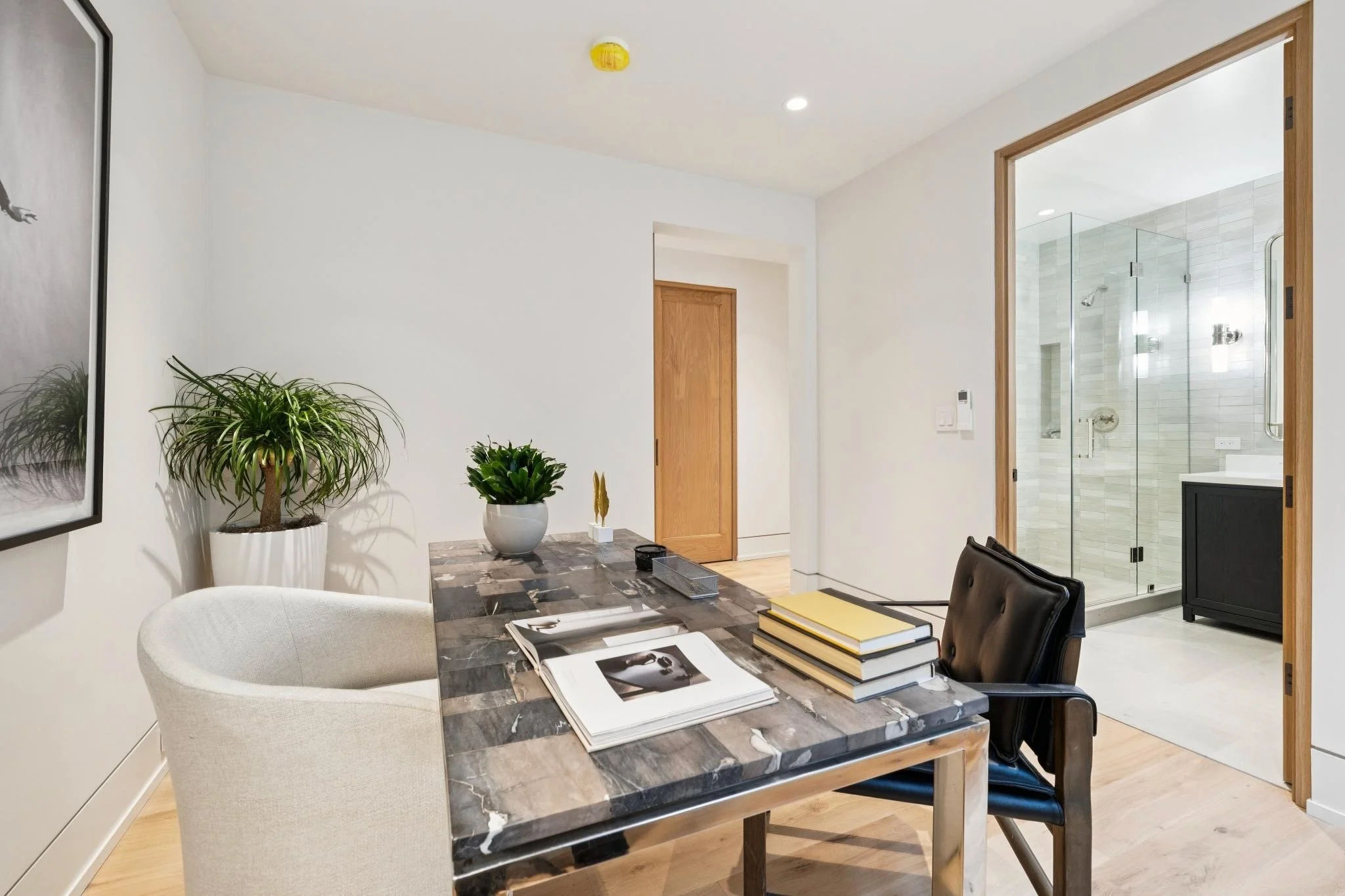 Modern interior room with a marble-topped dining table, black and white chairs, indoor plants, and a view into a bathroom with a glass shower enclosure.