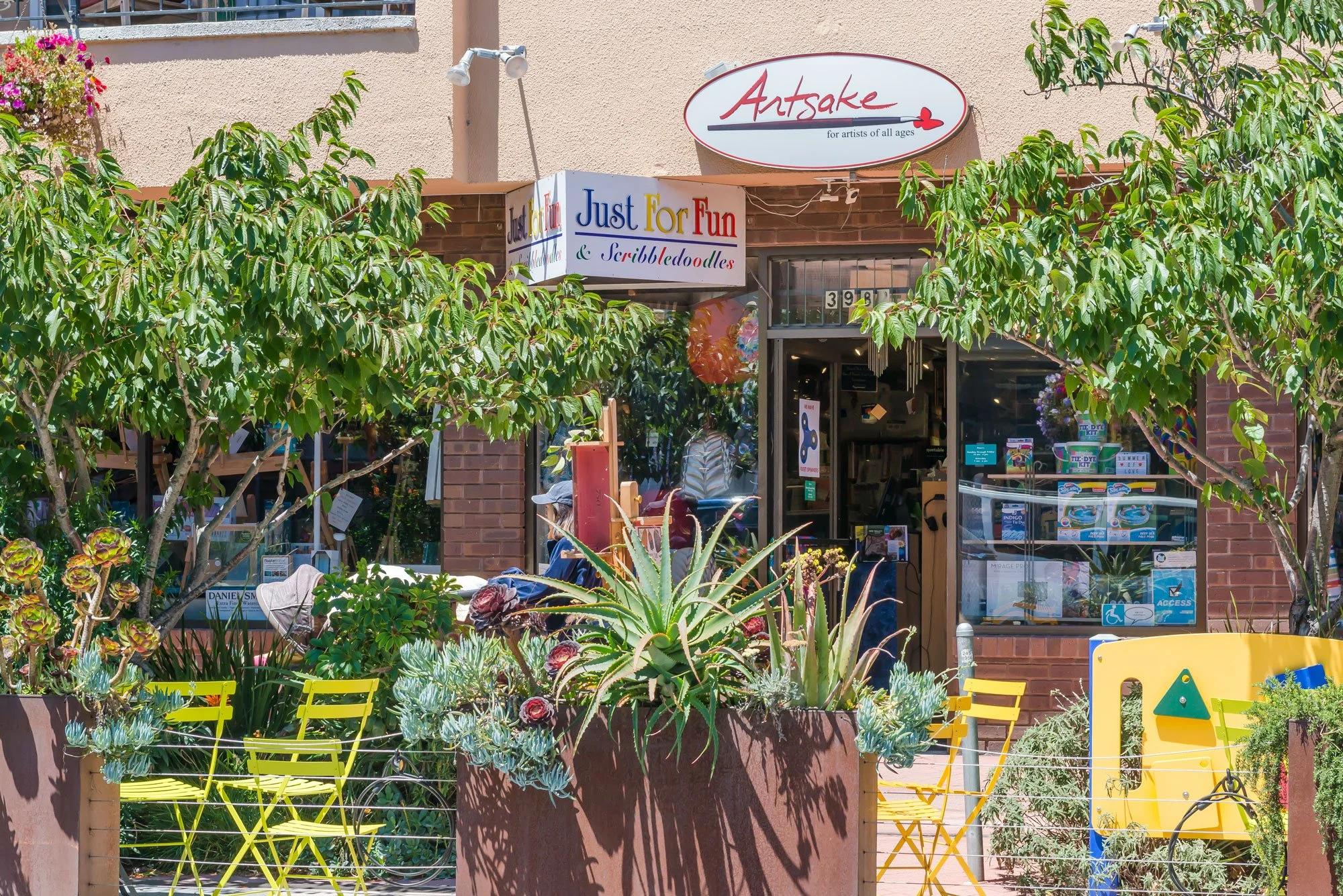 A storefront with green trees and plants outside, yellow chairs, and signs for an art store and a fun and doodles shop.