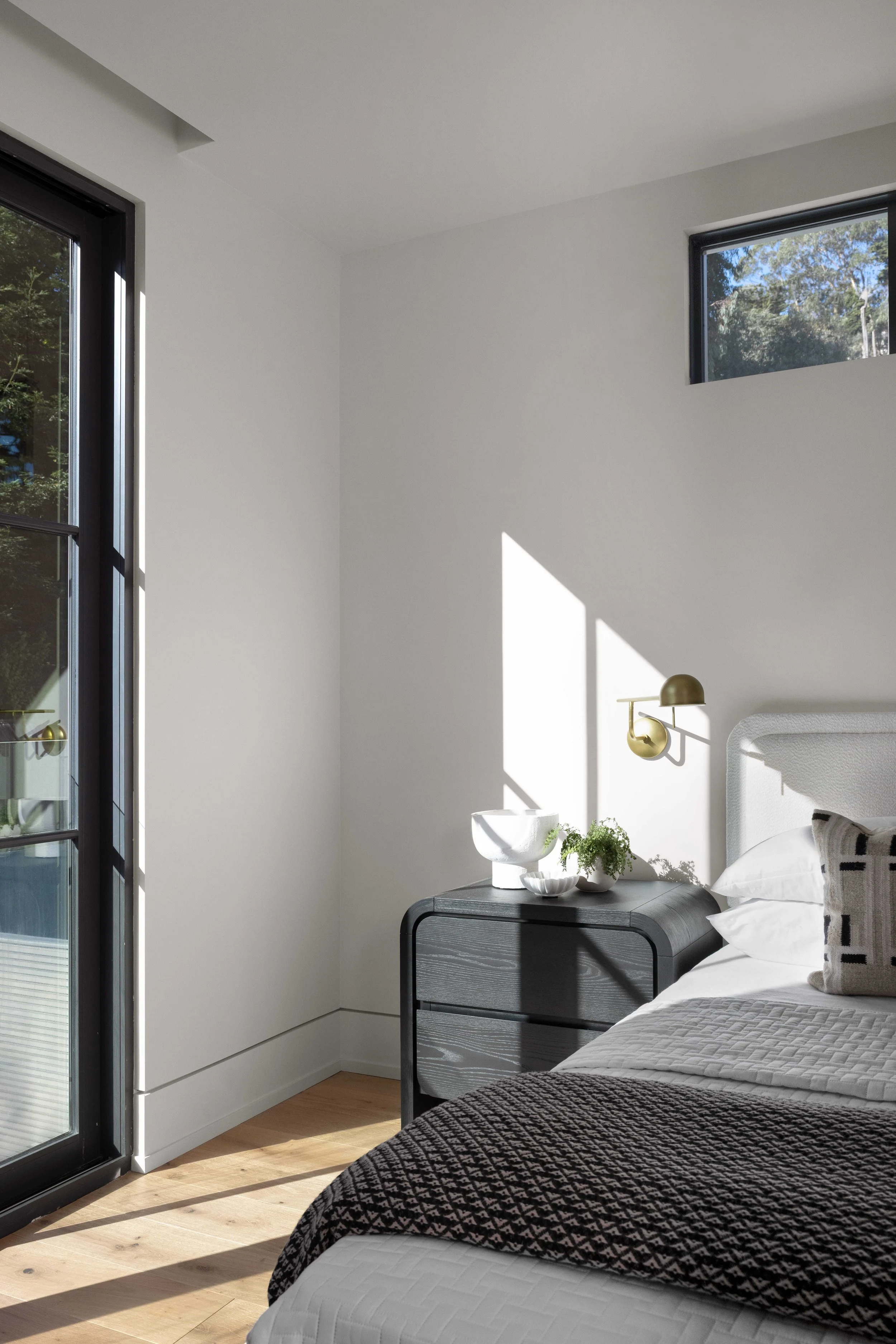 A bedroom with a white bed, black nightstand, wall-mounted lamp, and window letting in sunlight.
