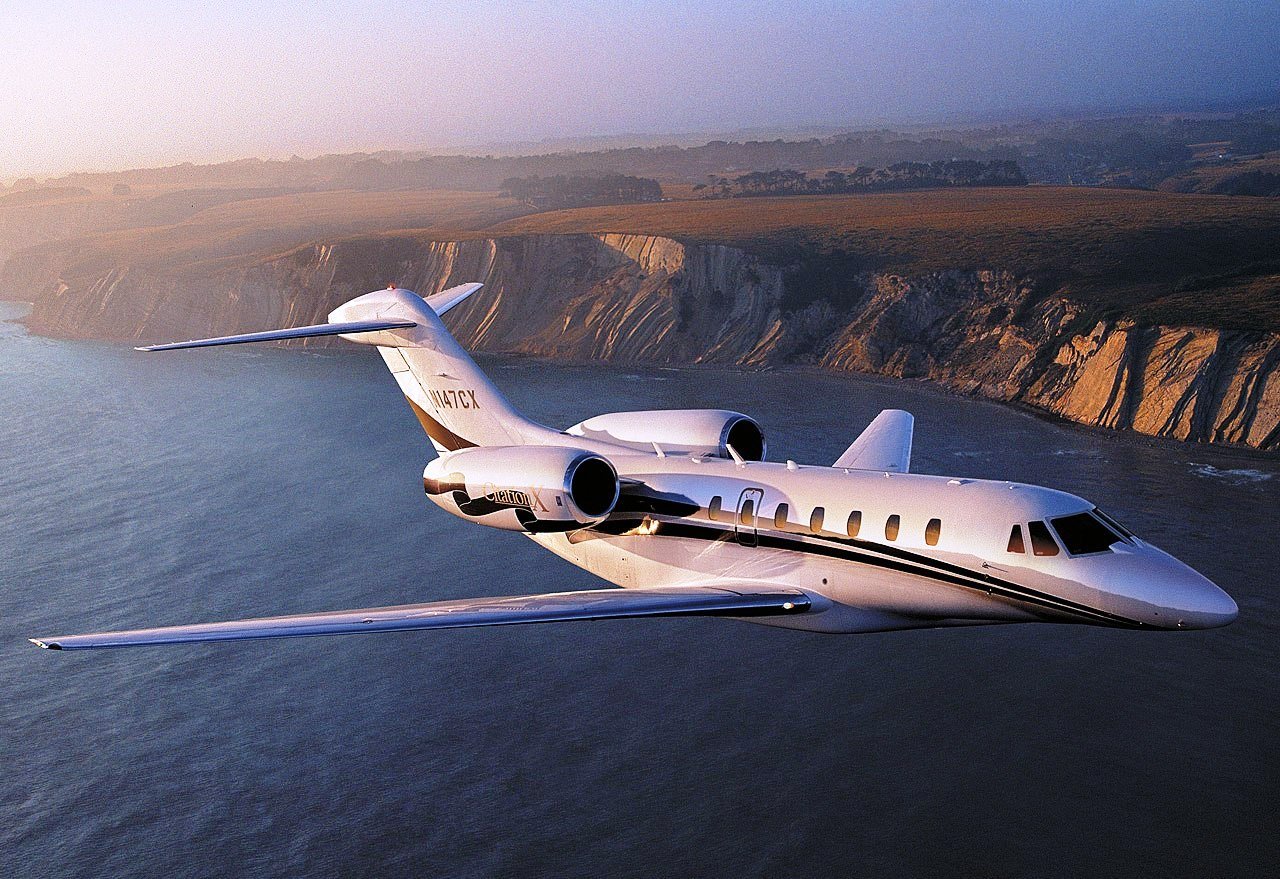 A private jet flying over a body of water with cliffs and hillside in the background during sunset.