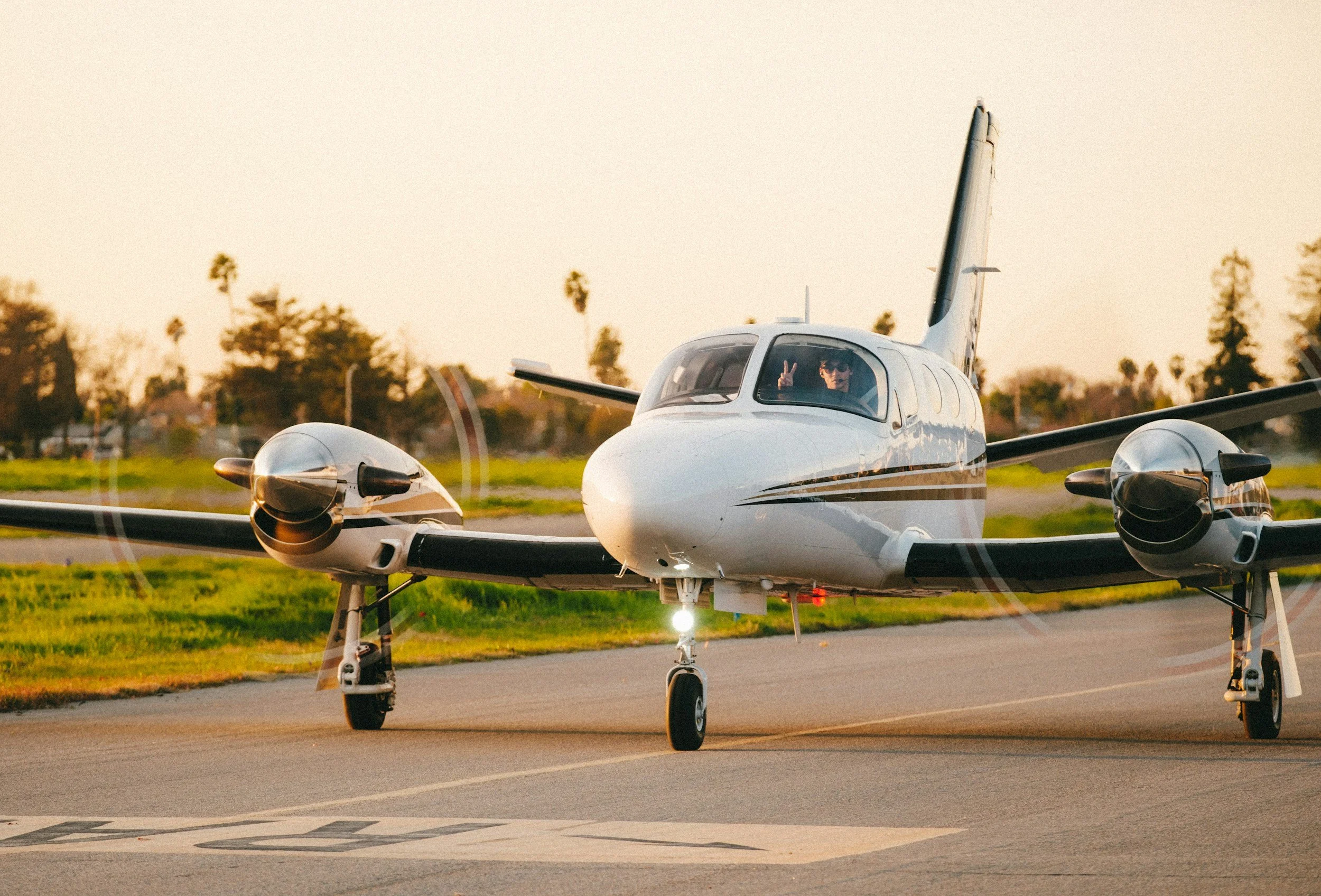 A Cessna Conquest prop jet is taxiing on the runway during sunset with two people inside, one of whom is waving peace signs.