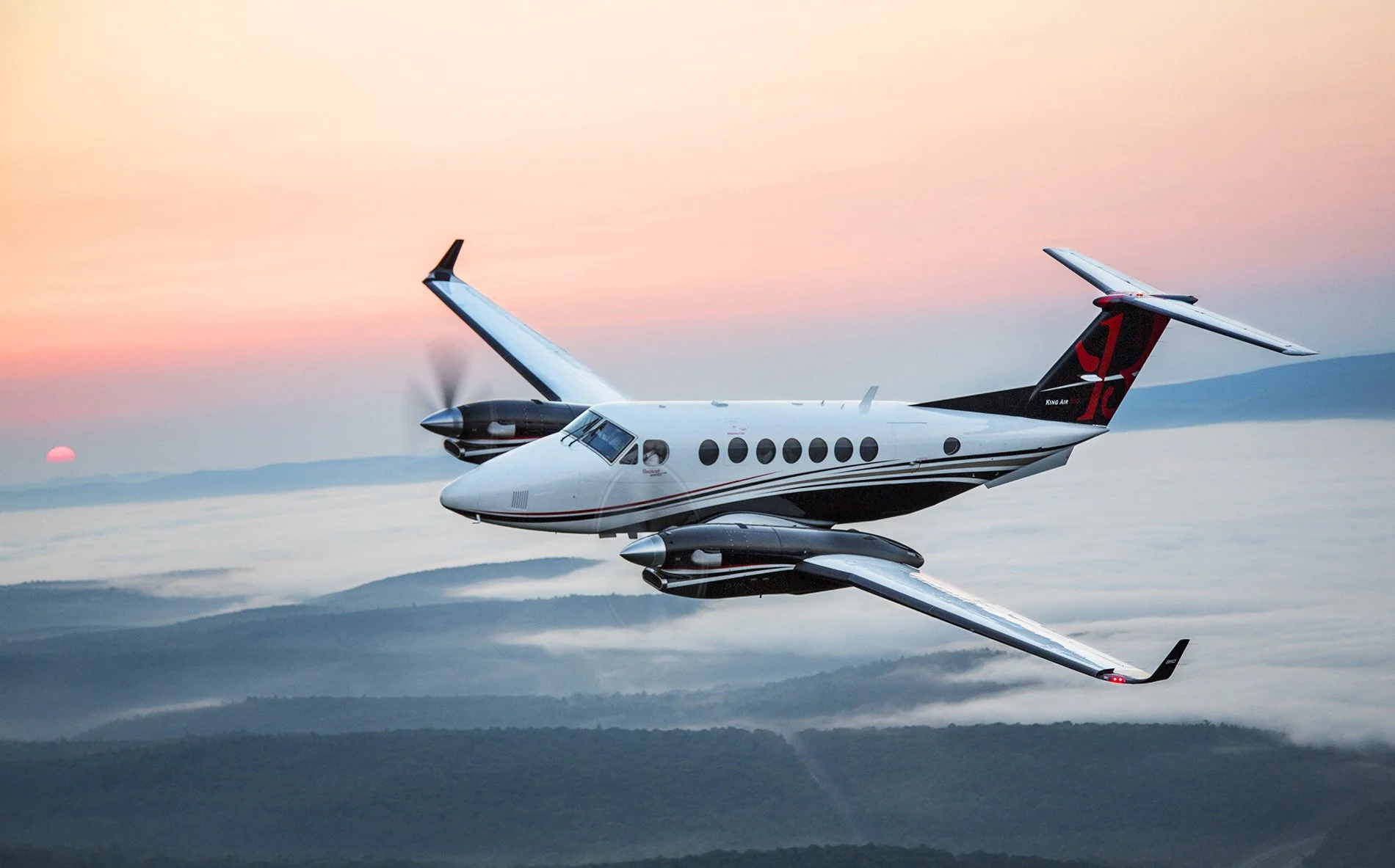 A twin-engine turboprop airplane flying above the clouds at sunset with mountains in the background.