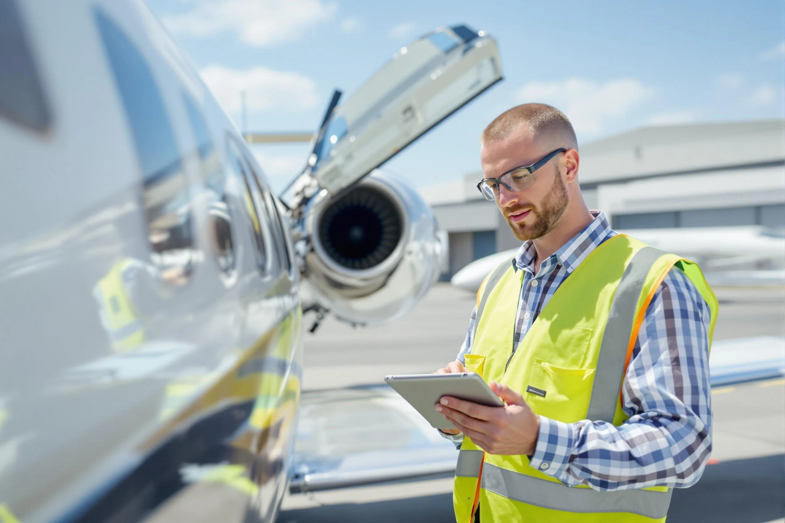 A man with glasses and a beard wearing a yellow safety vest and checked shirt is inspecting an airplane at an airport. He is holding a tablet and looking at the aircraft from the side. The airplane is a small private jet with the open door visible in the background. The sky is partly cloudy.