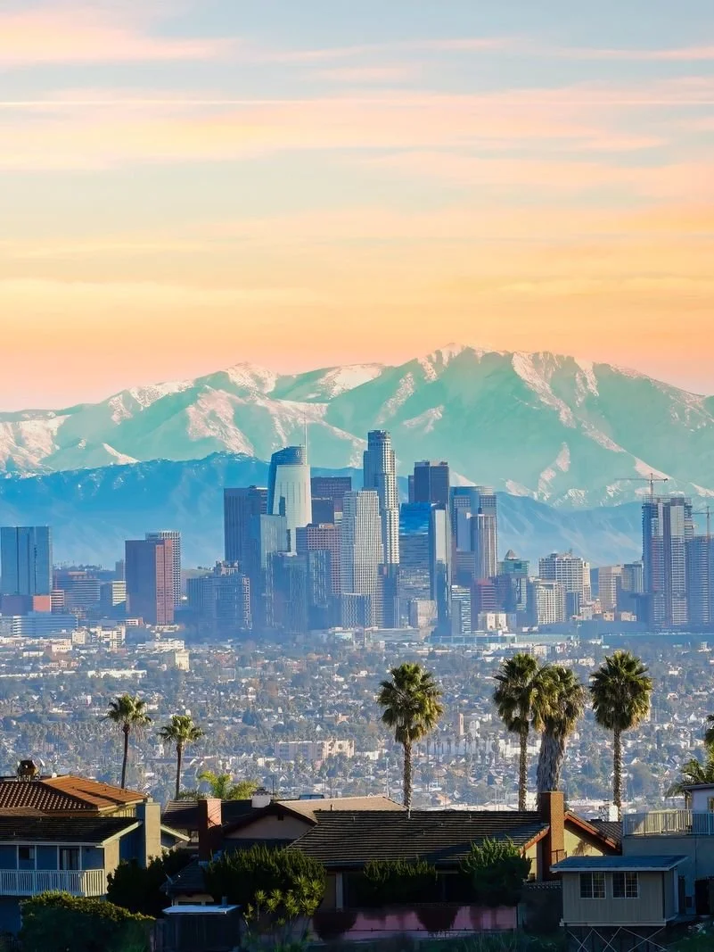 Sunset over downtown Los Angeles with the city skyline, palm trees in the foreground, and snow-capped mountains in the background.